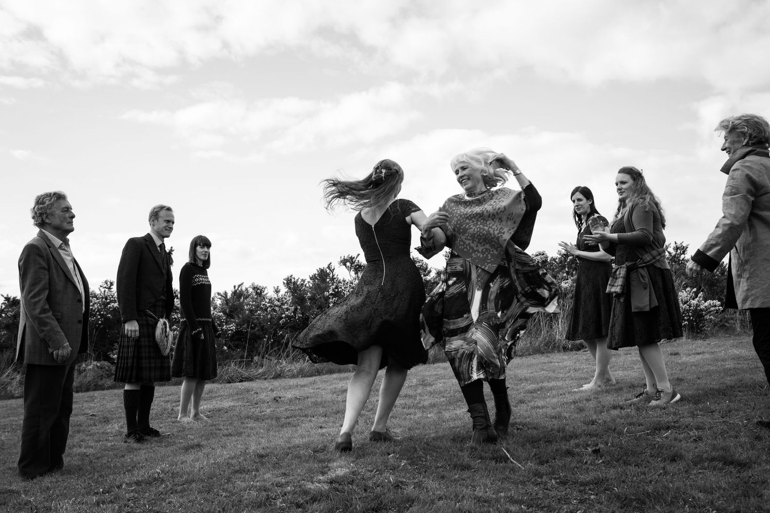 A vintage black and white photograph shows people dancing outdoors on a grassy hillside in a lively, energetic scene.
