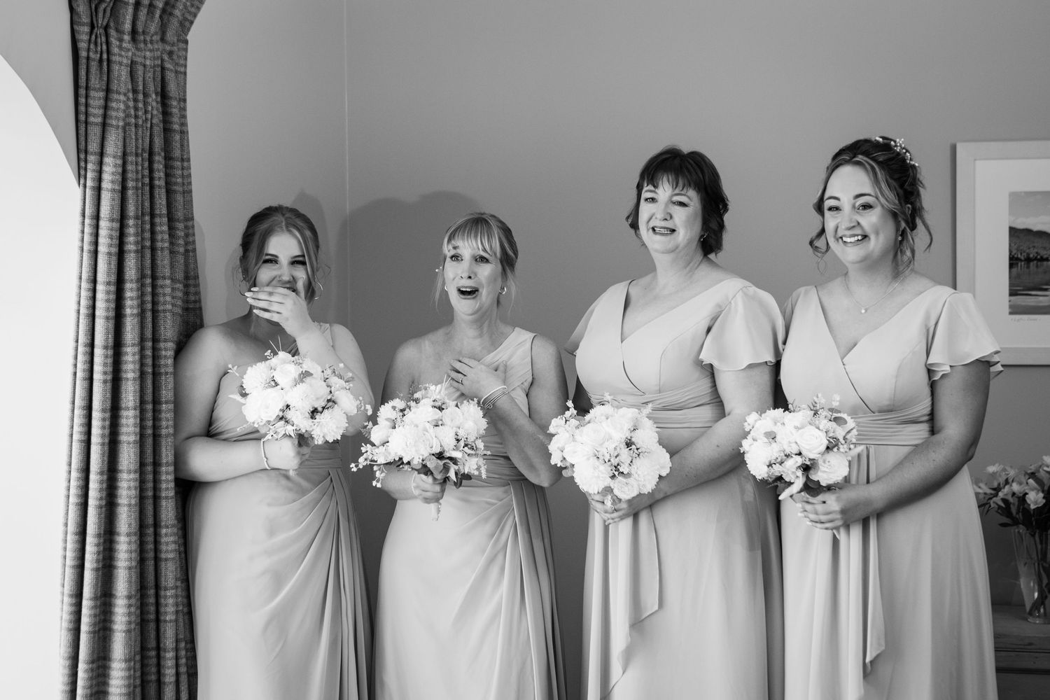 A row of bridesmaids in white dresses holding bouquets pose together against a light background.