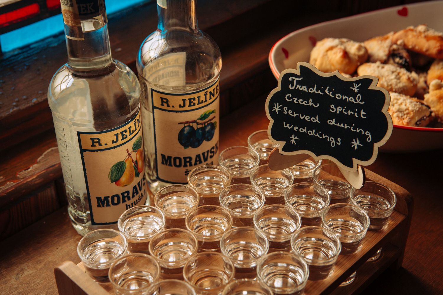 Shot glasses filled with clear liquor are arranged on a rustic wooden bar counter next to bottles and snacks.