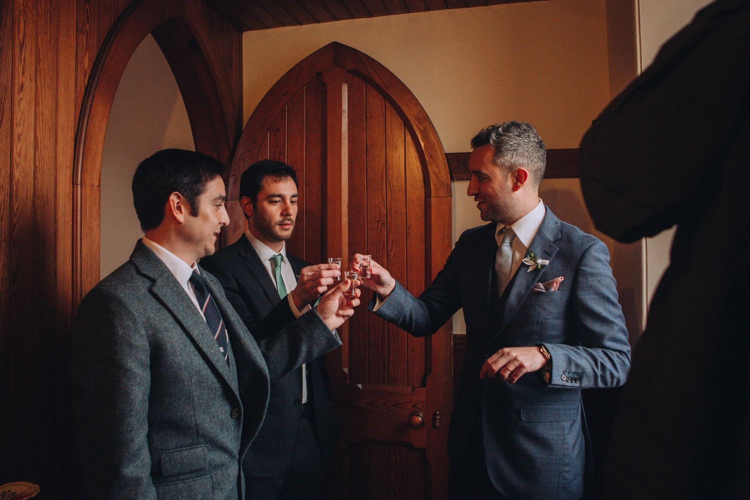 Wedding party members in gray suits share candid moments and laughter in an elegant wood-paneled venue.