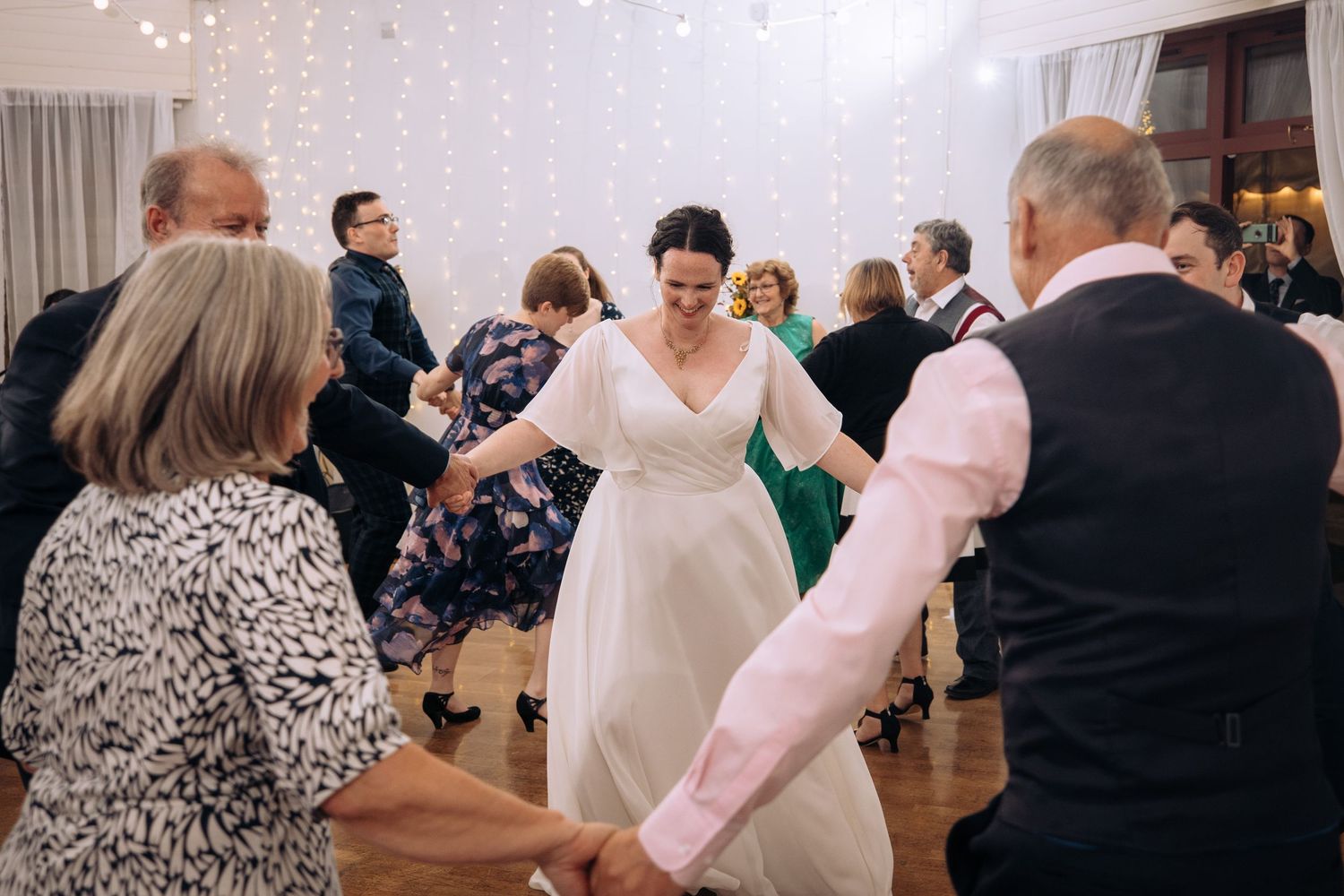 Wedding guests dance together on a wooden dance floor under twinkling string lights at an indoor reception.