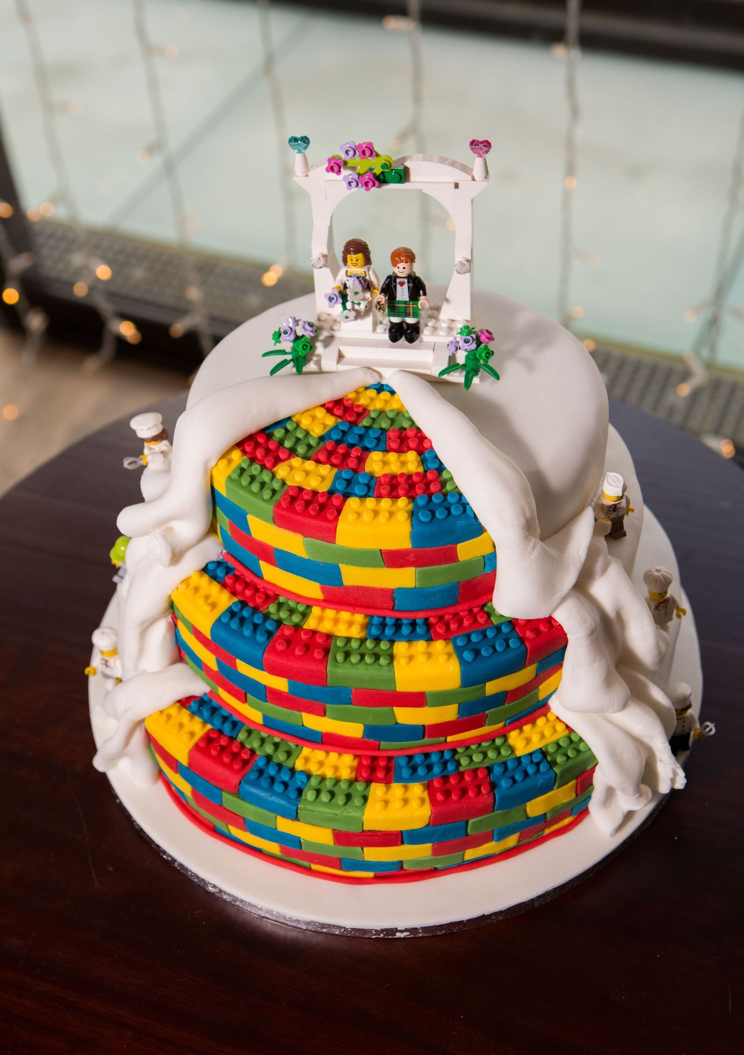 A wedding cake decorated with colorful LEGO bricks and topped with LEGO minifigures under a white arch.
