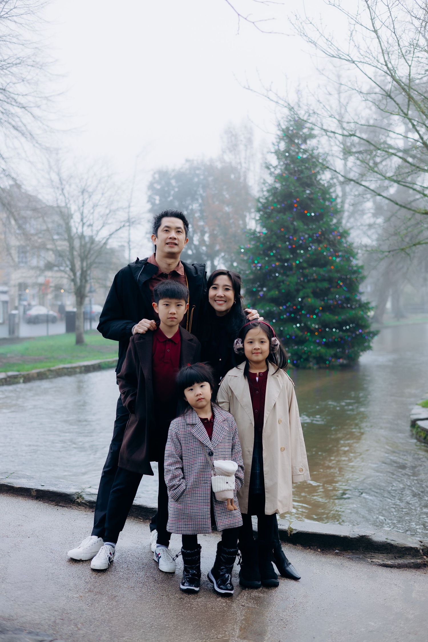 A family stands together near a decorated Christmas tree on a misty winter day by the water.