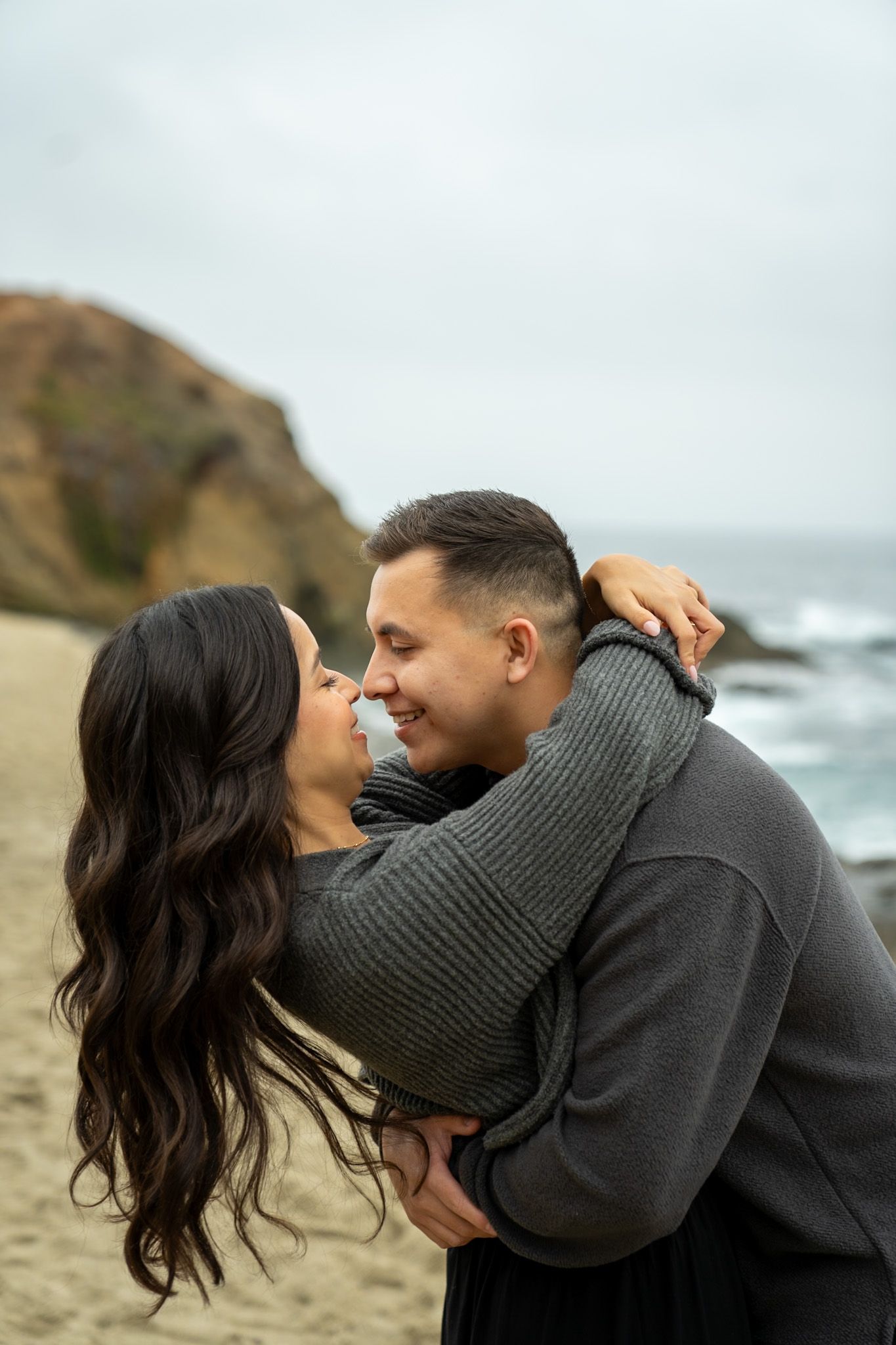 A romantic couple shares intimate moments at a scenic coastal location wearing cozy sweaters on an overcast day.