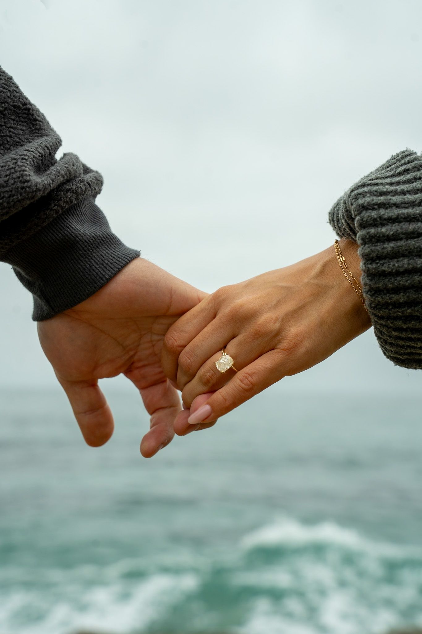 Two hands with wedding rings reach toward each other against a blurry ocean backdrop, symbolizing love and commitment.