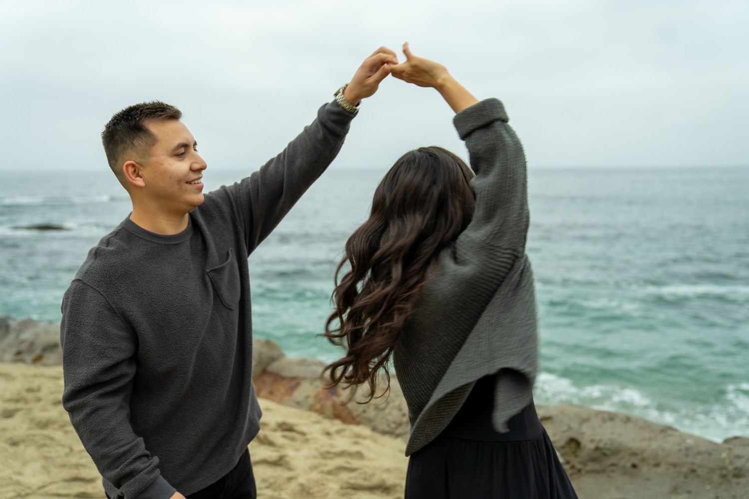 Couple embraces on rocky coastal cliff with ocean waves in background.