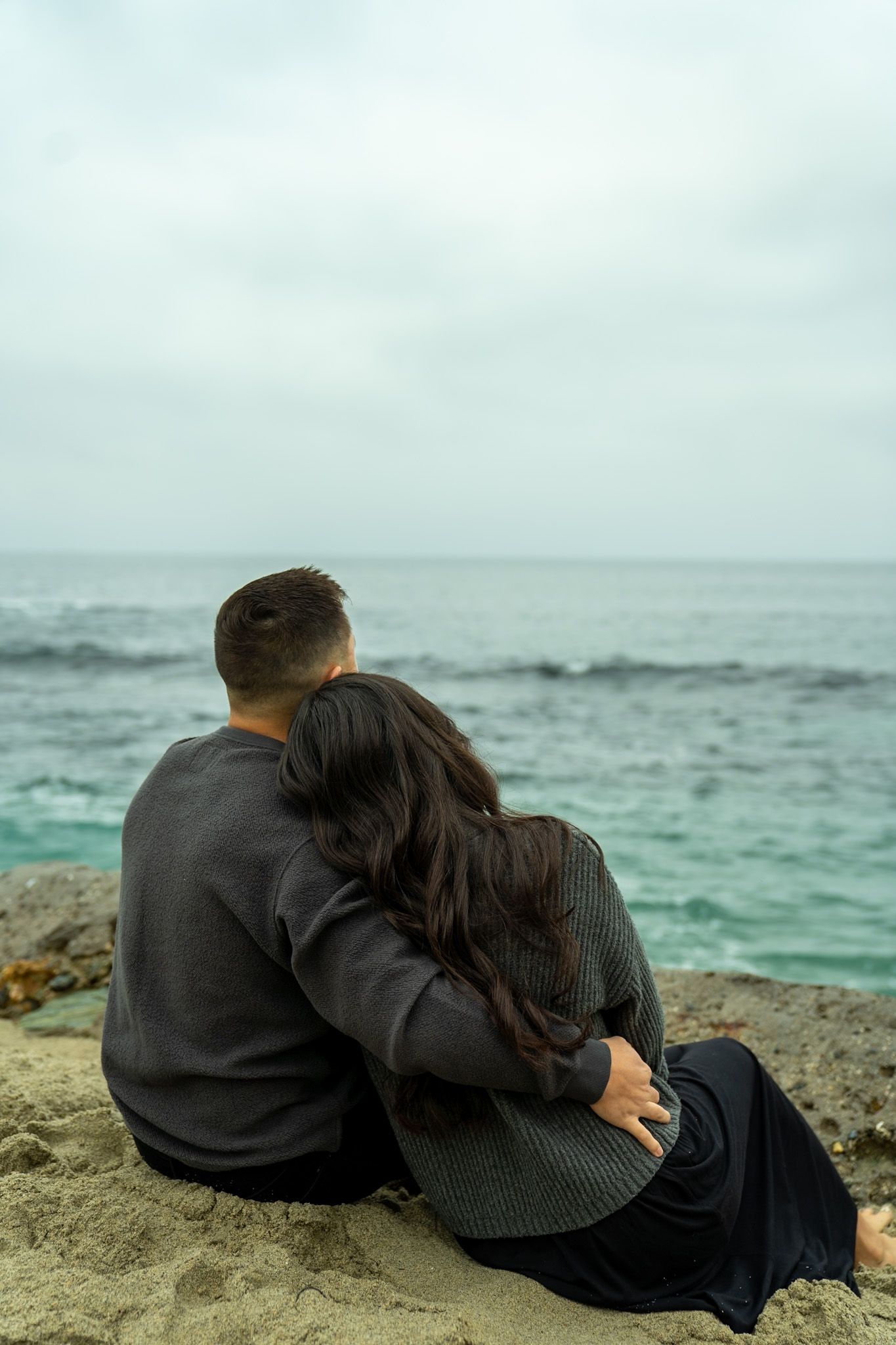 A couple sits together on rocky coastline overlooking turquoise ocean waters on an overcast day.