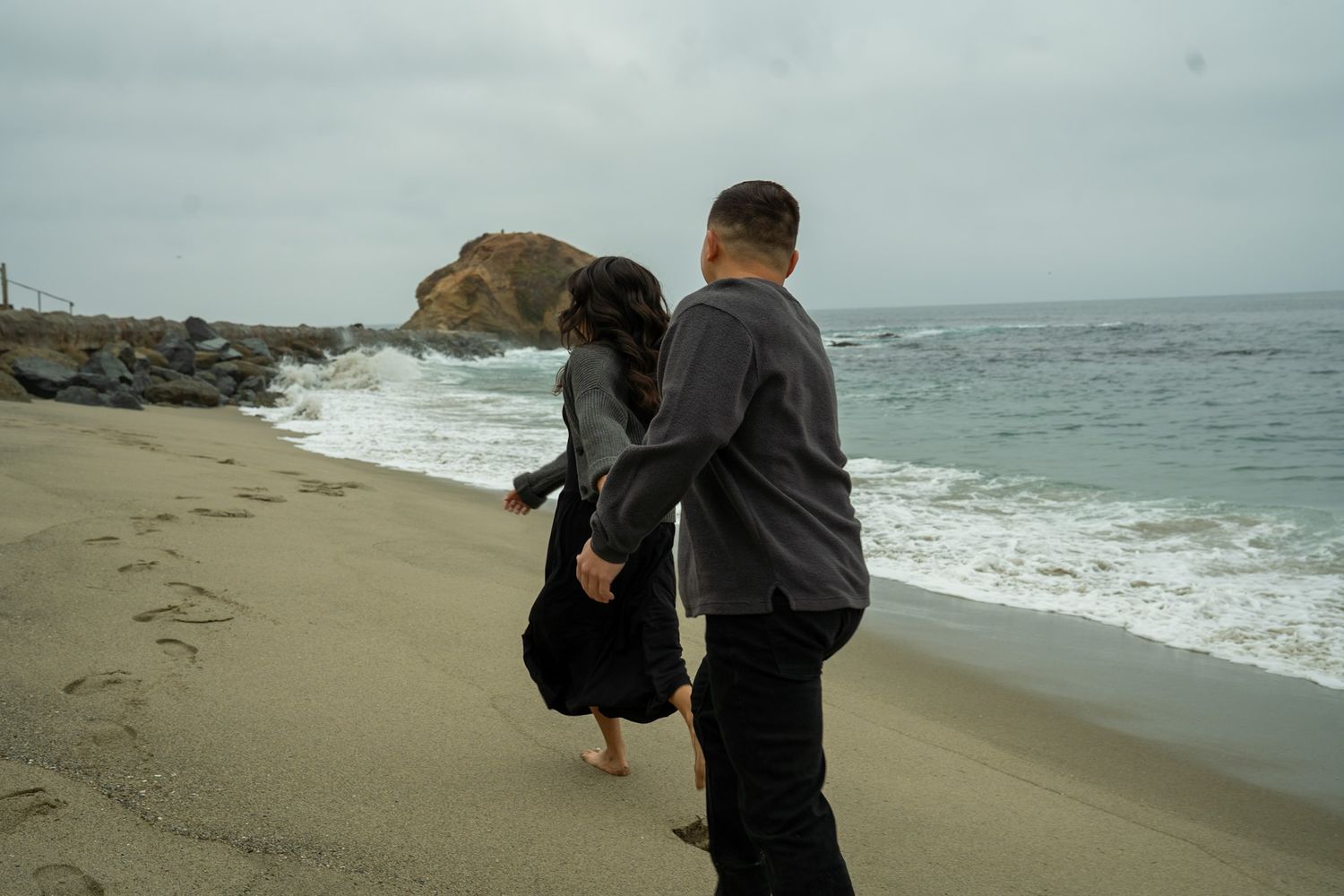 Two people walk along a misty beach shoreline while waves crash against the rocky coast on an overcast day.