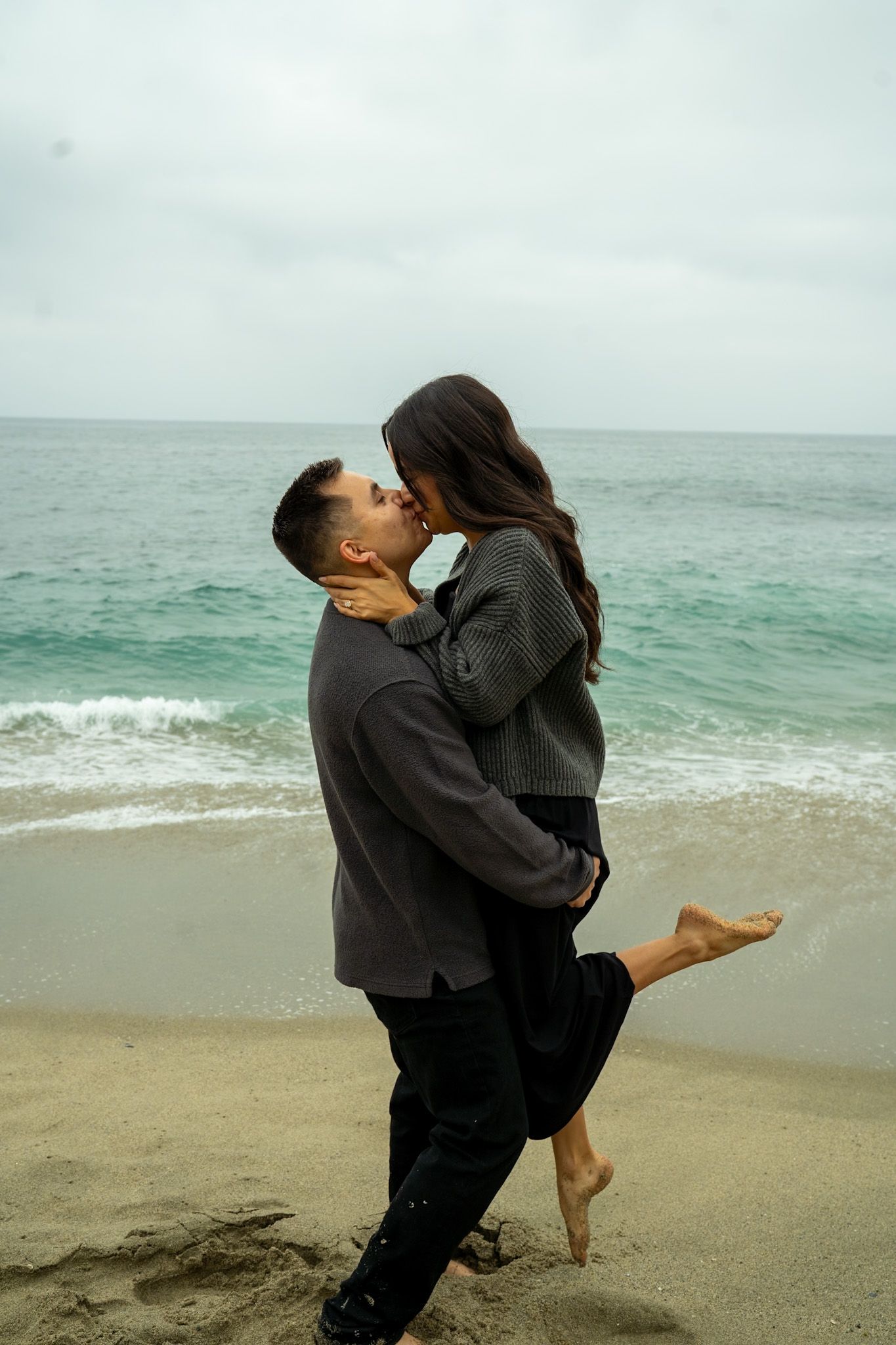 A romantic couple embraces on a beach with crashing waves and cloudy skies in the background.