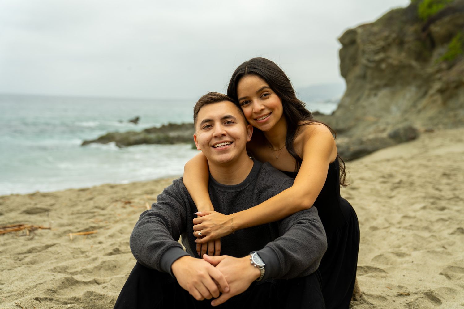 A couple embraces on a scenic beach with rocky cliffs and ocean waves in the background.