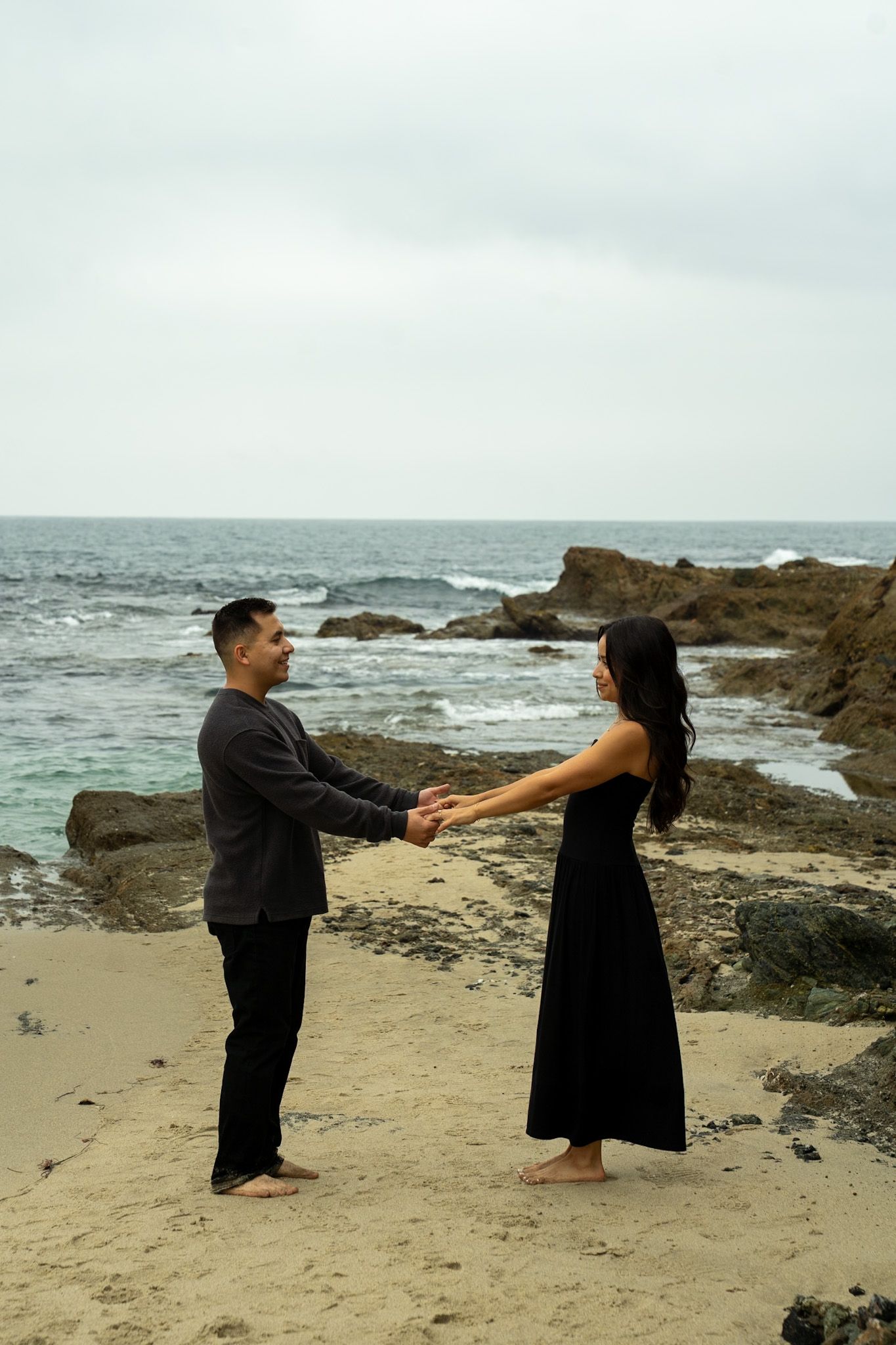 A couple holds hands while standing barefoot on a rocky beach with ocean waves and cliffs in the background.