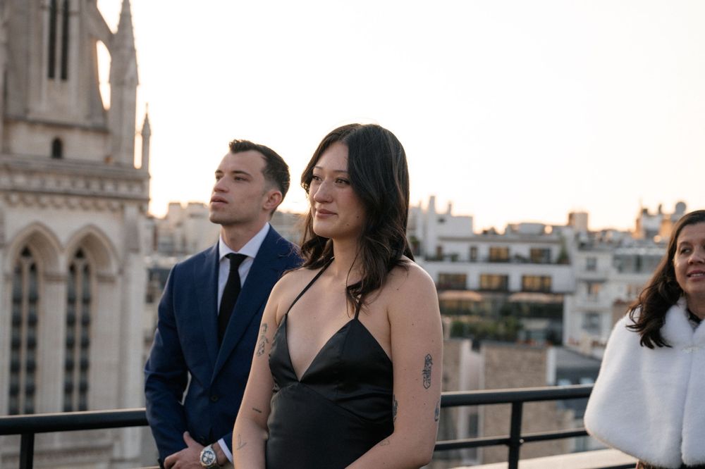 A group of well-dressed people gather on a rooftop terrace with a historic European cityscape visible in the background.