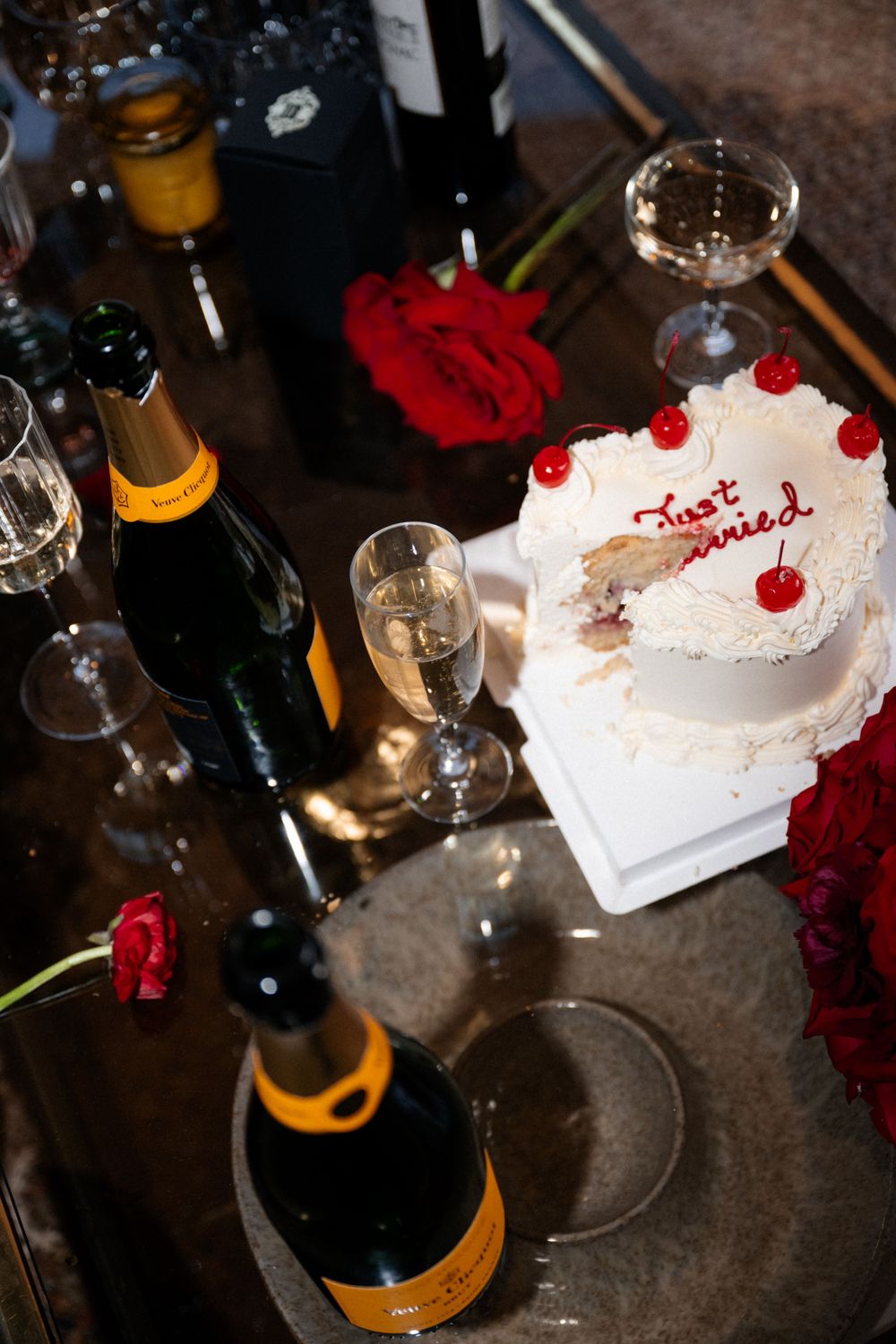A celebration setup with champagne bottles and a white cake topped with red cherries on a dark table.