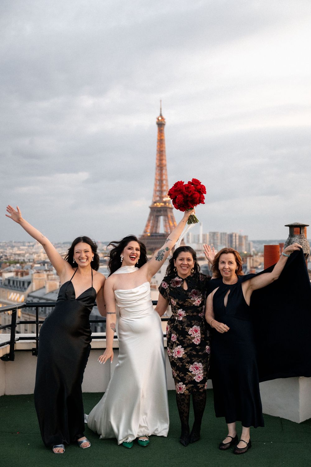 A group celebrates joyfully on a rooftop terrace with the Eiffel Tower visible in the Paris skyline at dusk.