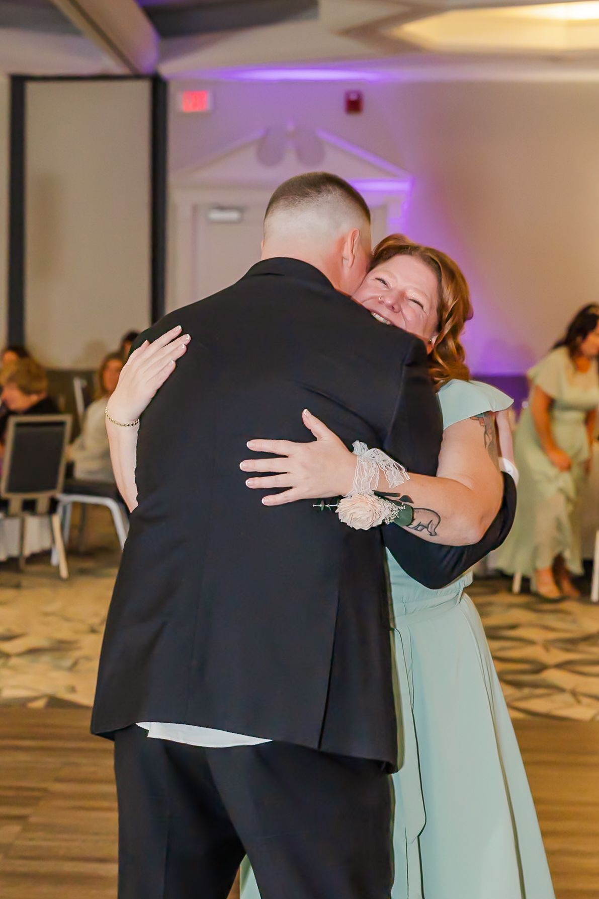 A couple in formal attire share an emotional embrace during what appears to be a wedding reception.