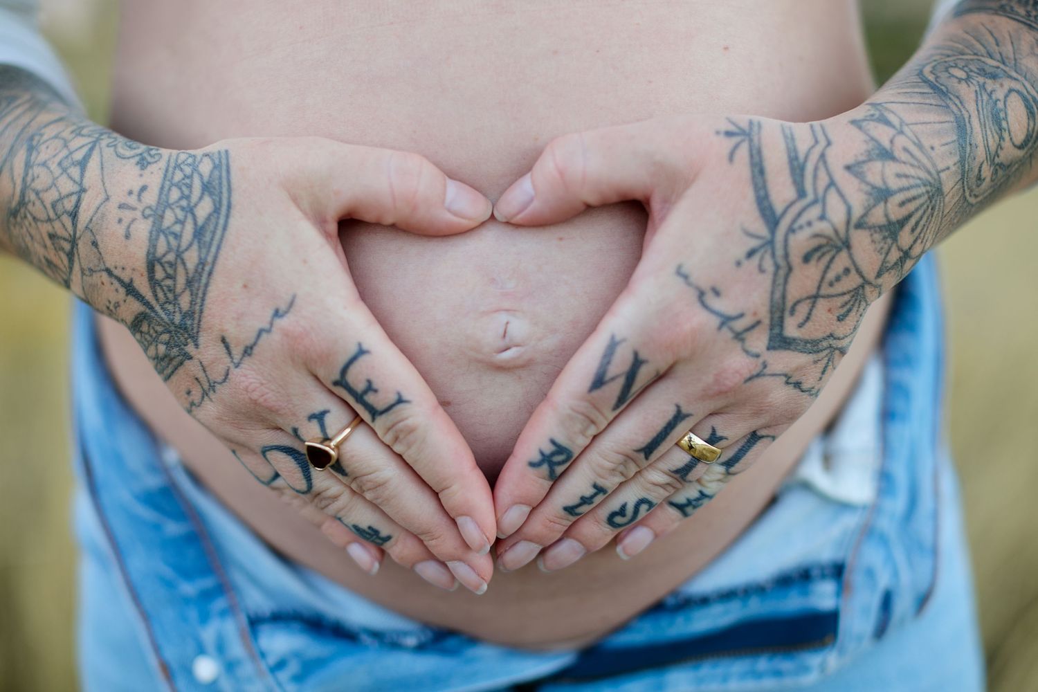 Tattooed hands form a heart shape over a pregnant belly in blue denim shorts.
