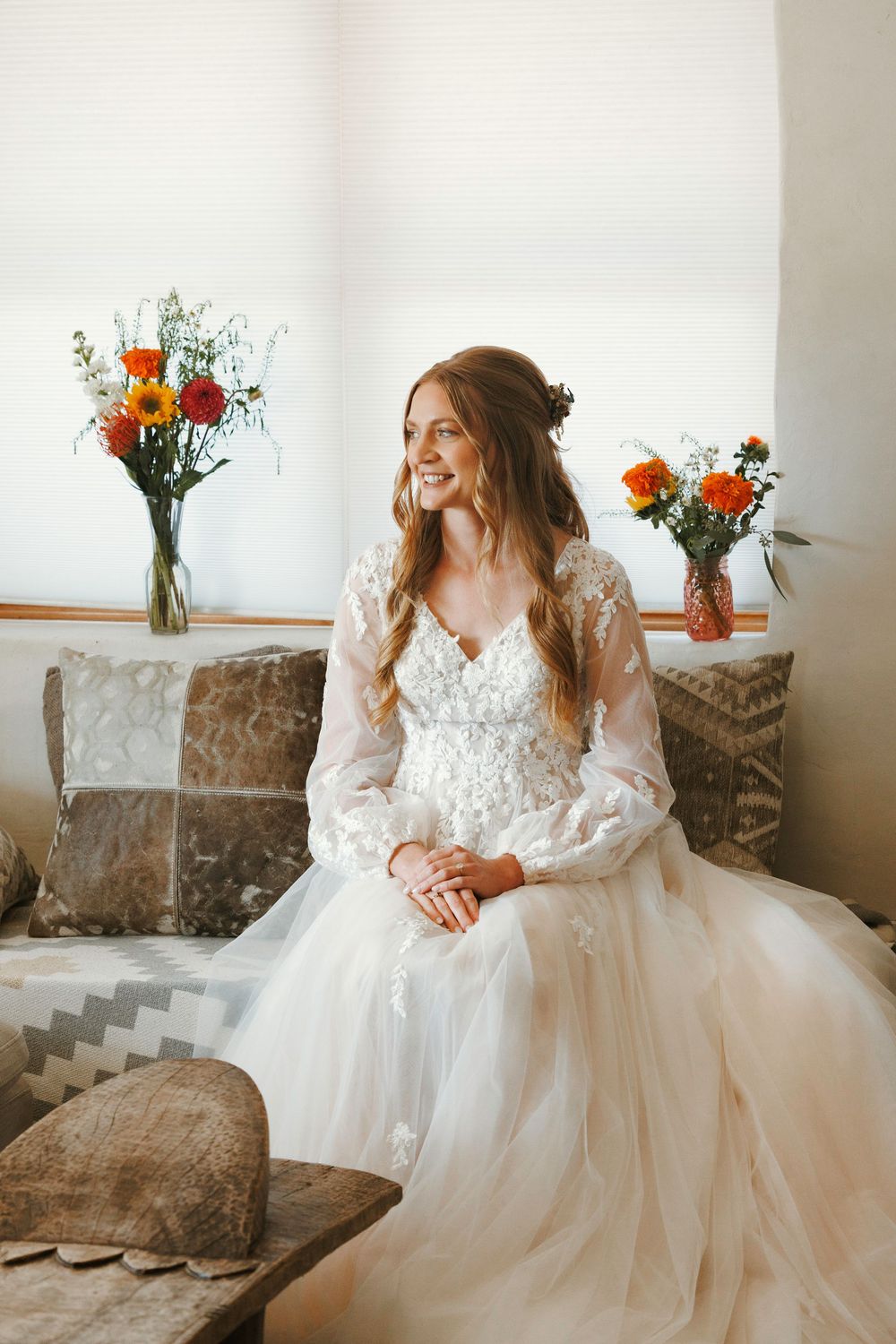 Smiling bride in lace wedding gown seated on couch with colorful flower arrangements nearby.