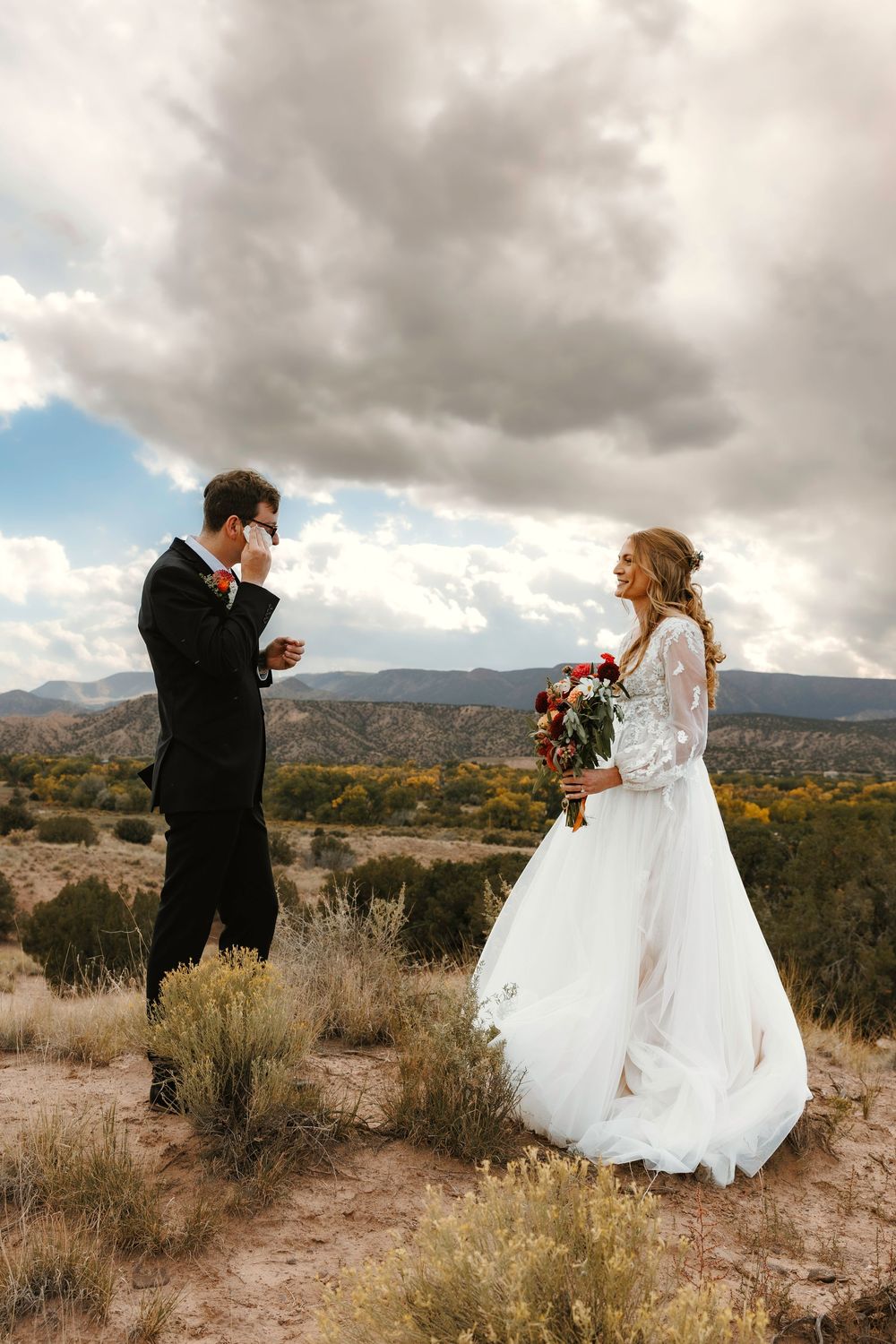 Groom wipes tears seeing bride in lace gown for first time in desert landscape.