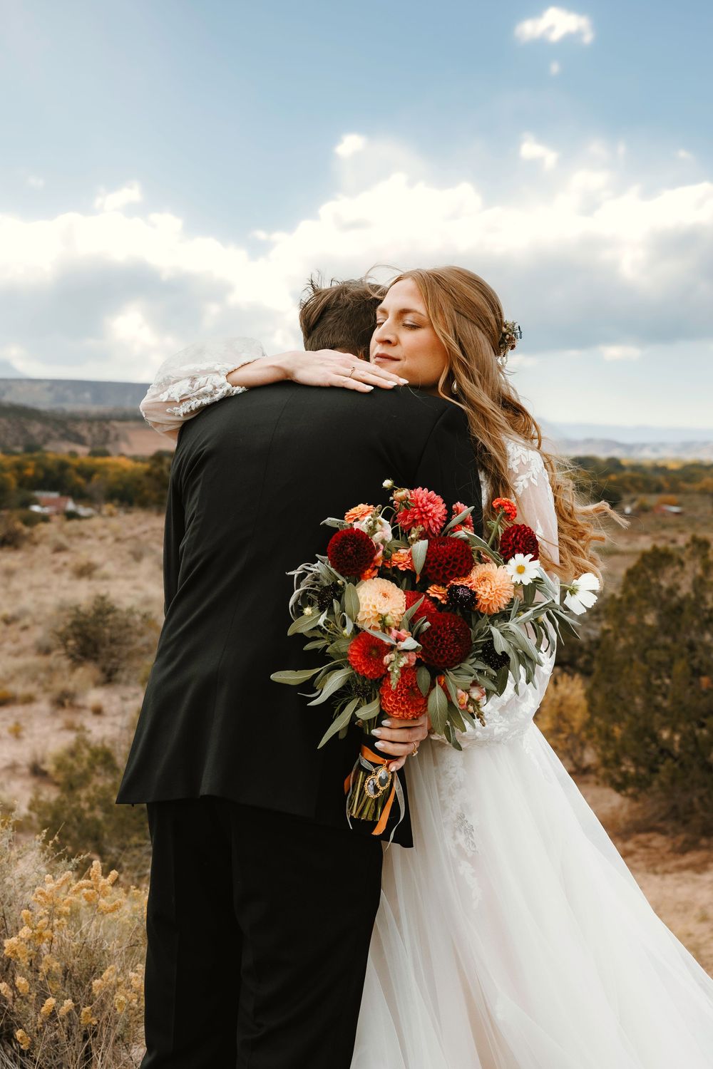 Bride holding red bouquet embraces groom with eyes closed in scenic desert setting.