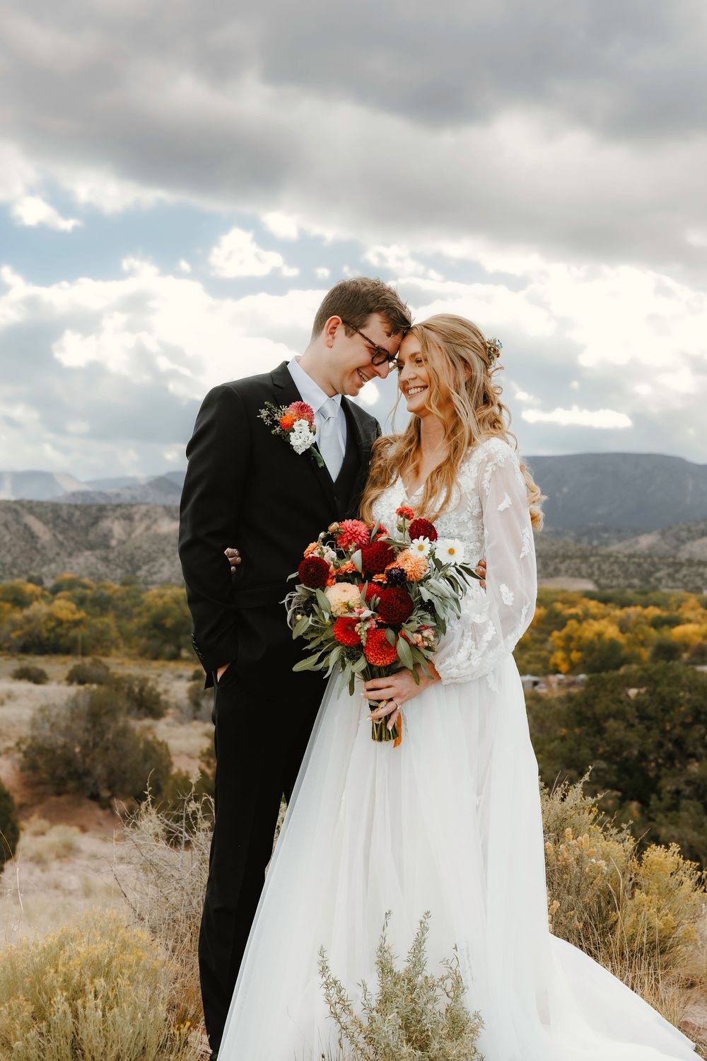 Bride and groom embracing outdoors with dramatic mountain landscape and cloudy sky behind them.