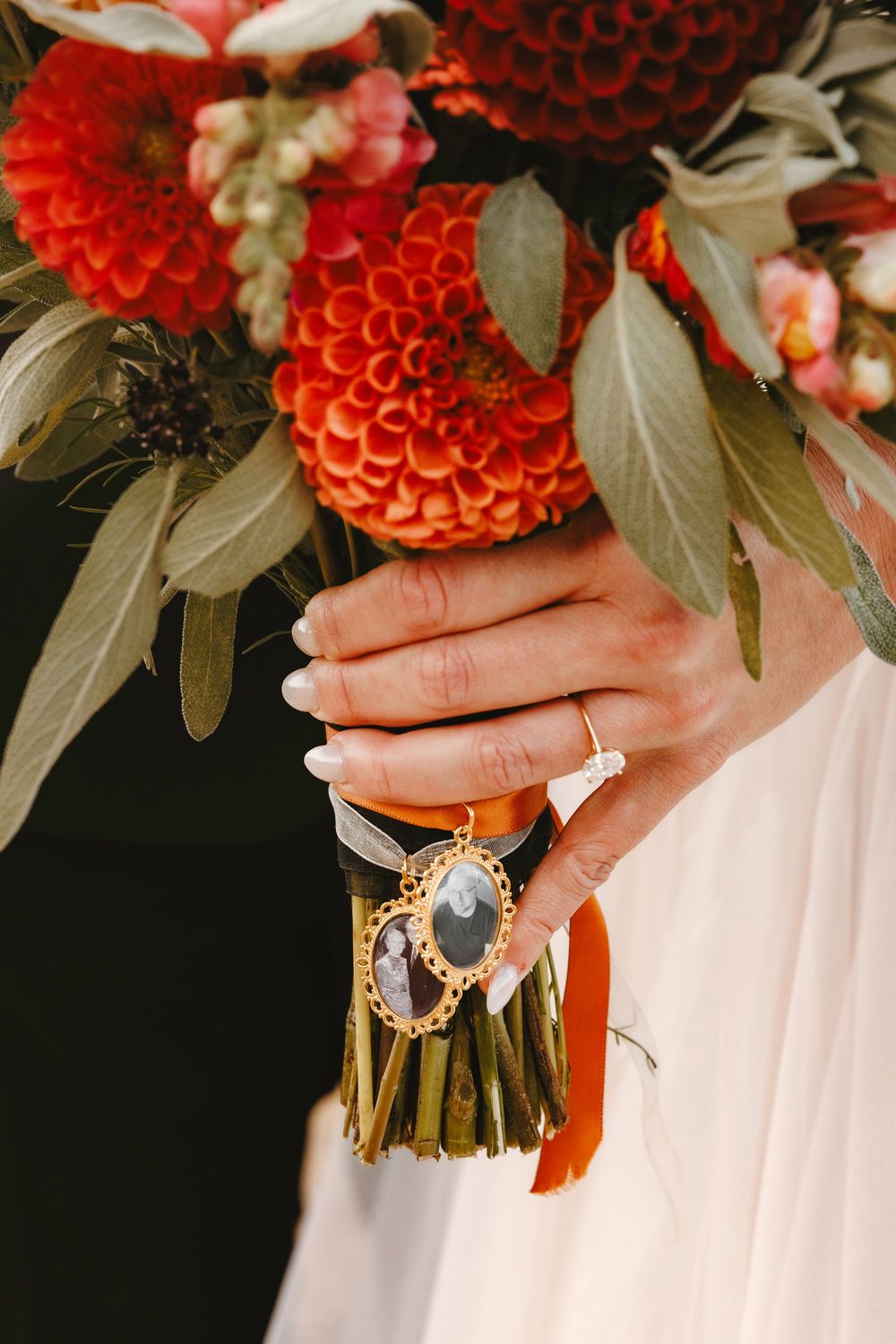 Bride holding red dahlia bouquet with gold memorial photo charm and white manicured nails.