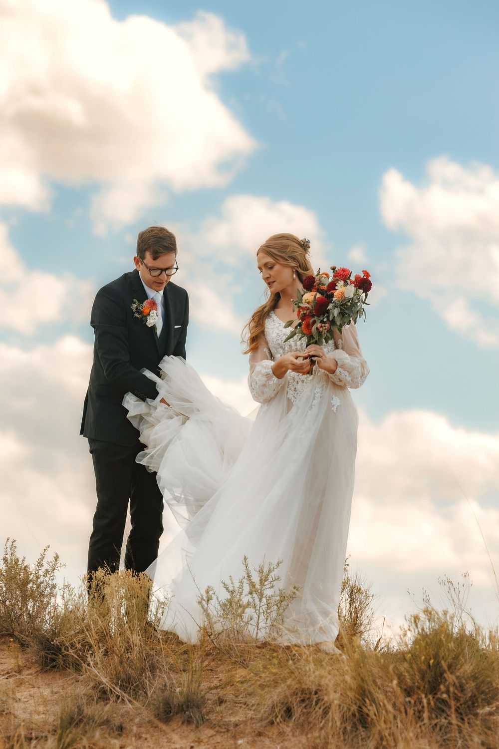 Groom helping bride with flowing white tulle dress on sandy hill under blue cloudy sky.