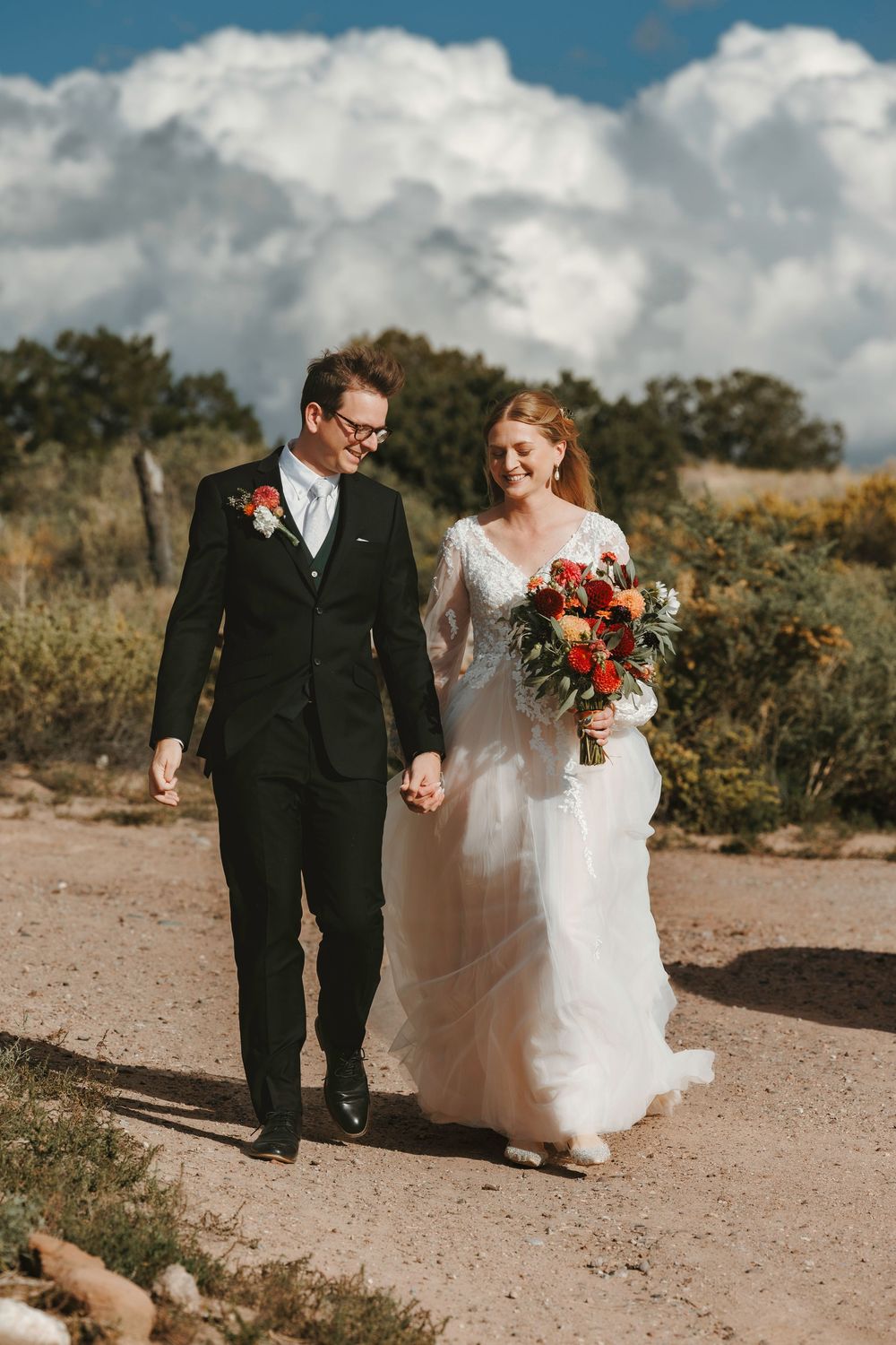 Smiling bride and groom holding hands walking outdoors with large white clouds in background.