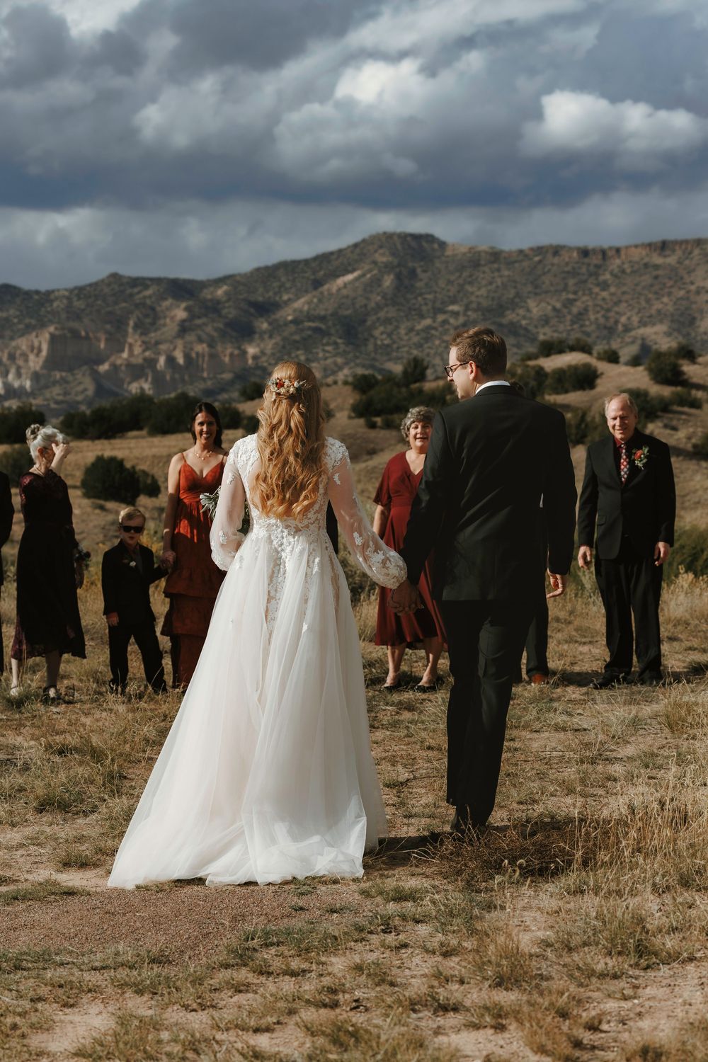 Bride and groom walking toward wedding party with rugged mountain backdrop under cloudy sky.