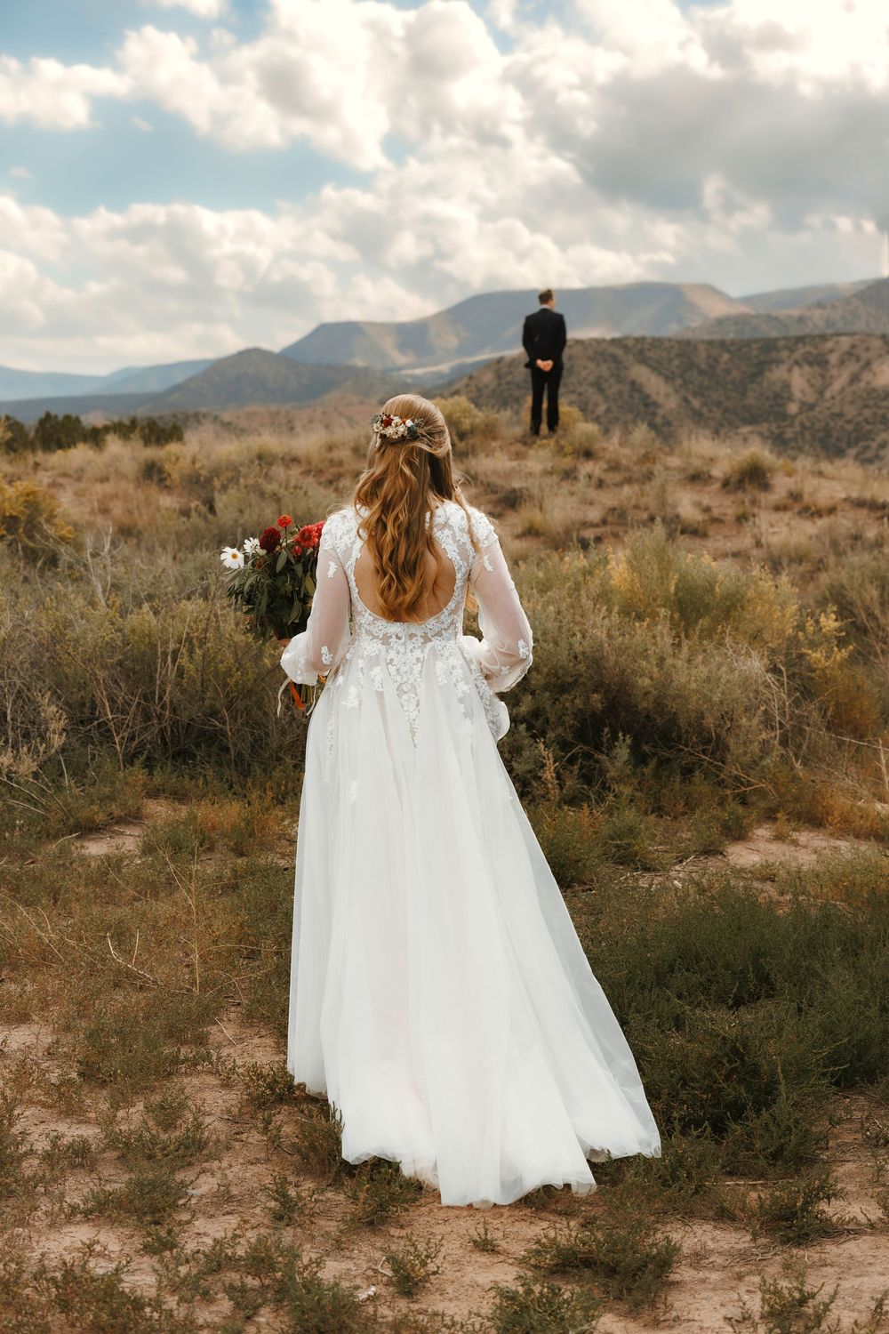 Bride in backless lace gown holding bouquet walks toward groom in desert landscape.