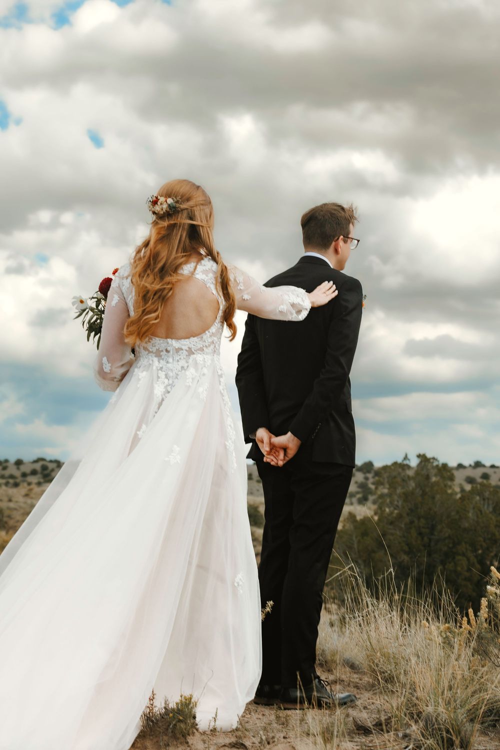 Bride and groom standing back-to-back holding hands under dramatic cloudy sky in desert.
