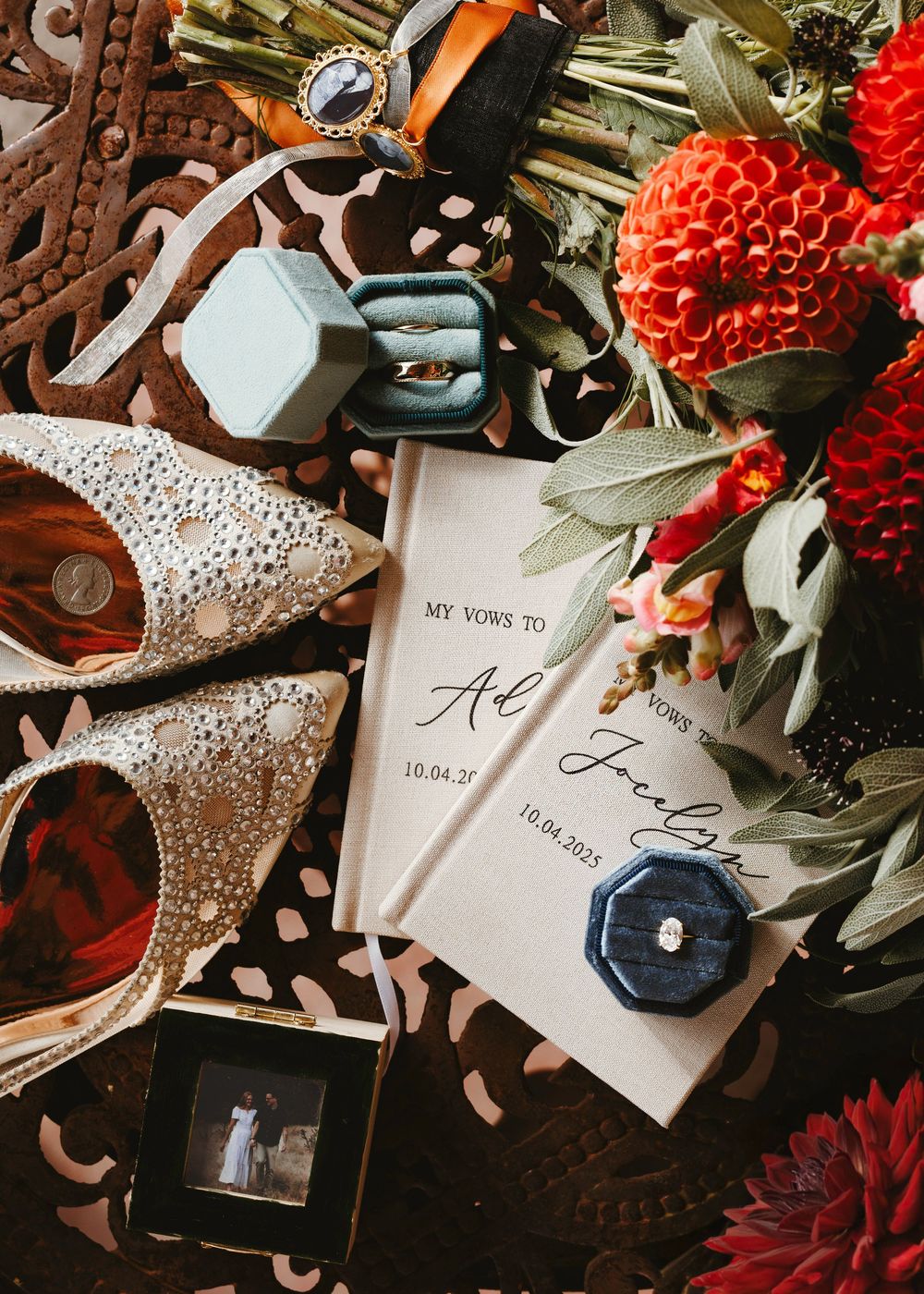 Wedding flat lay with vow books, engagement rings in velvet boxes, bridal heels, and red dahlia bouquet.