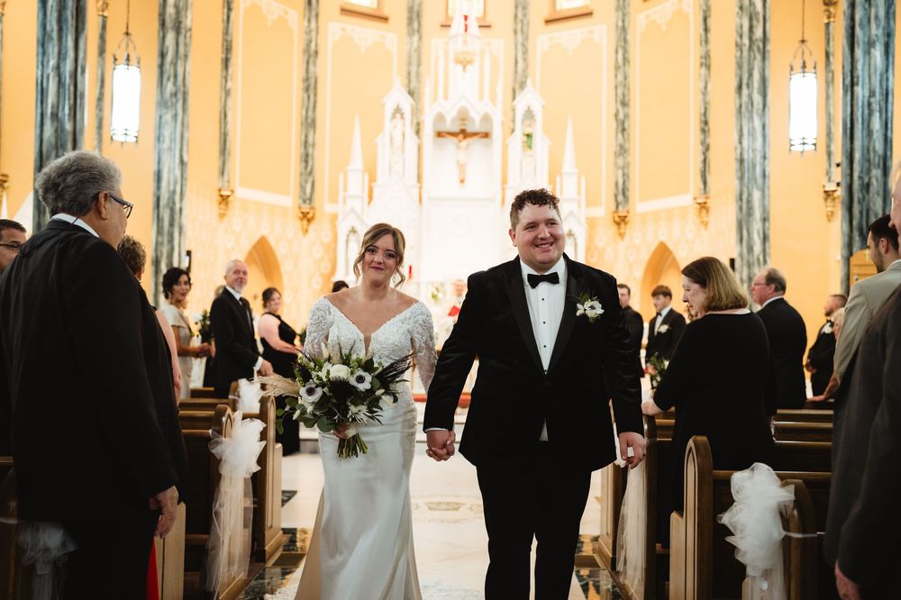 Wedding party walks down church aisle during ceremony in elegant cathedral with ornate architecture and stained glass windows.