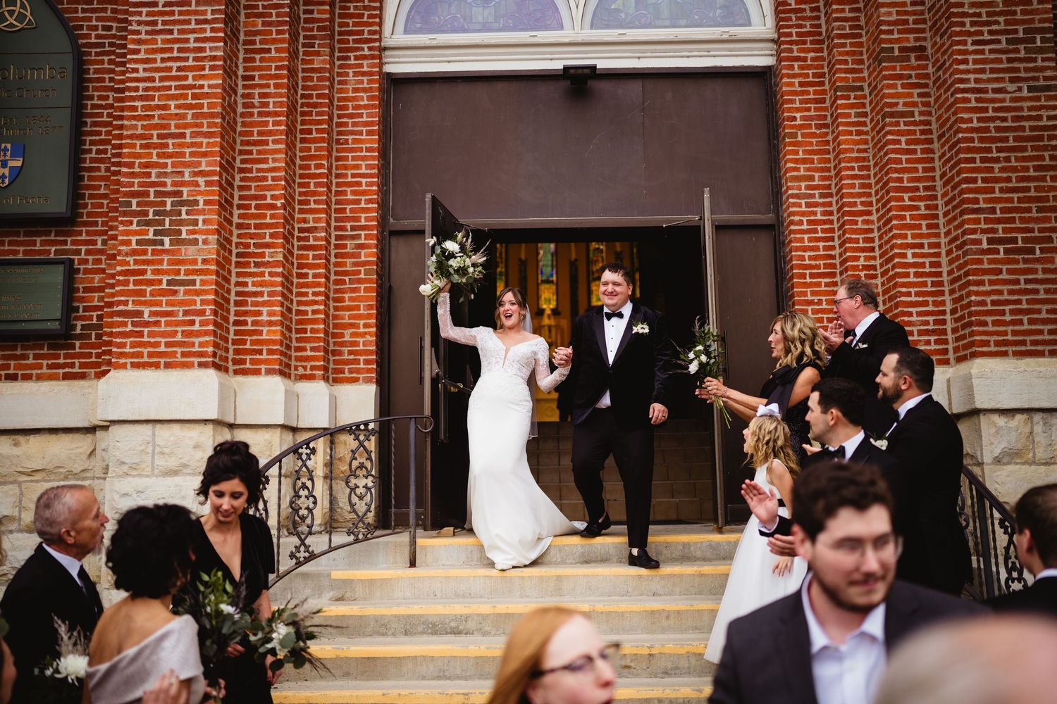 A newlywed couple exits a red brick church as guests celebrate with bubbles on the steps.
