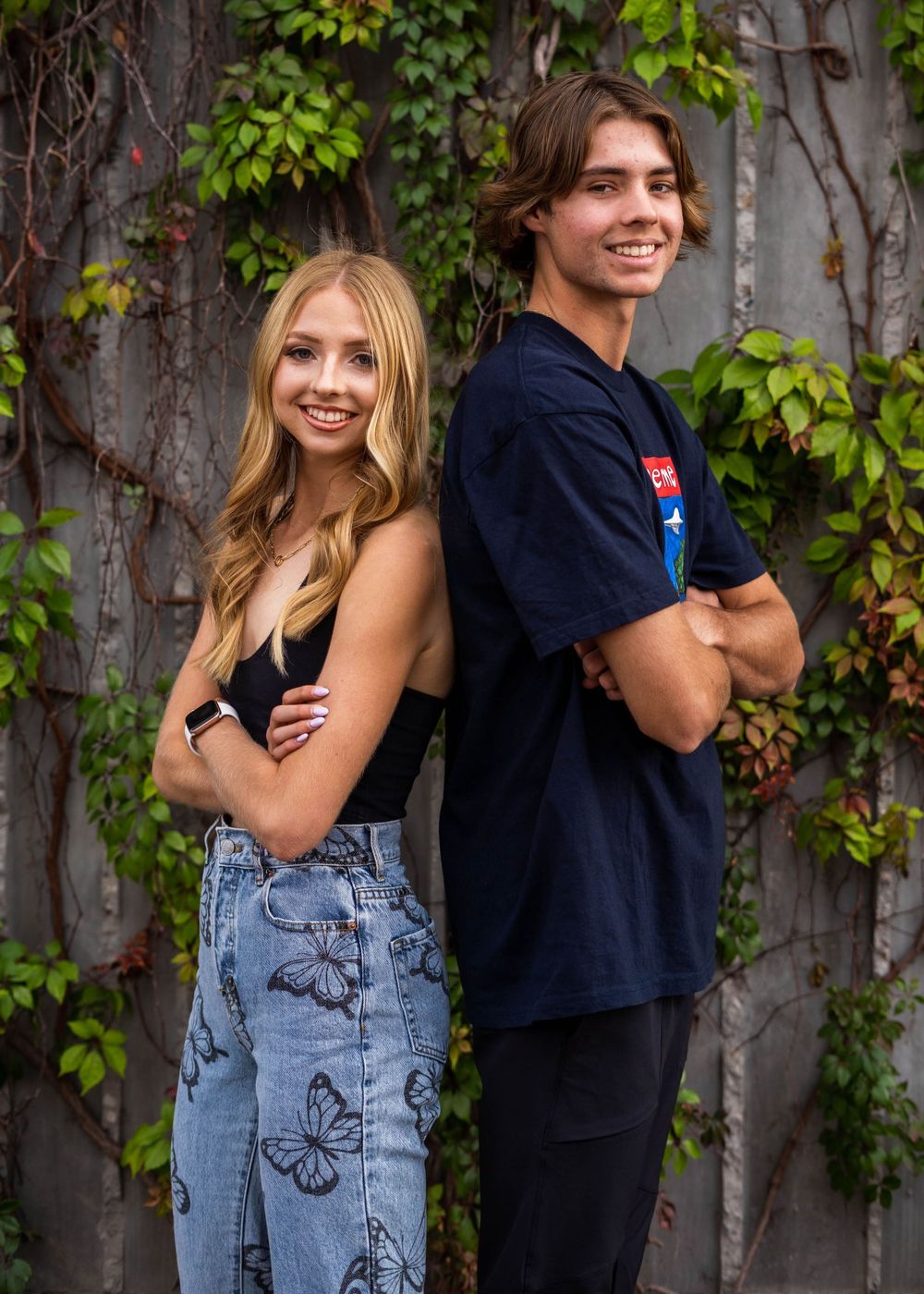 Two siblings pose back to back against a vine-covered wall in casual outfits.