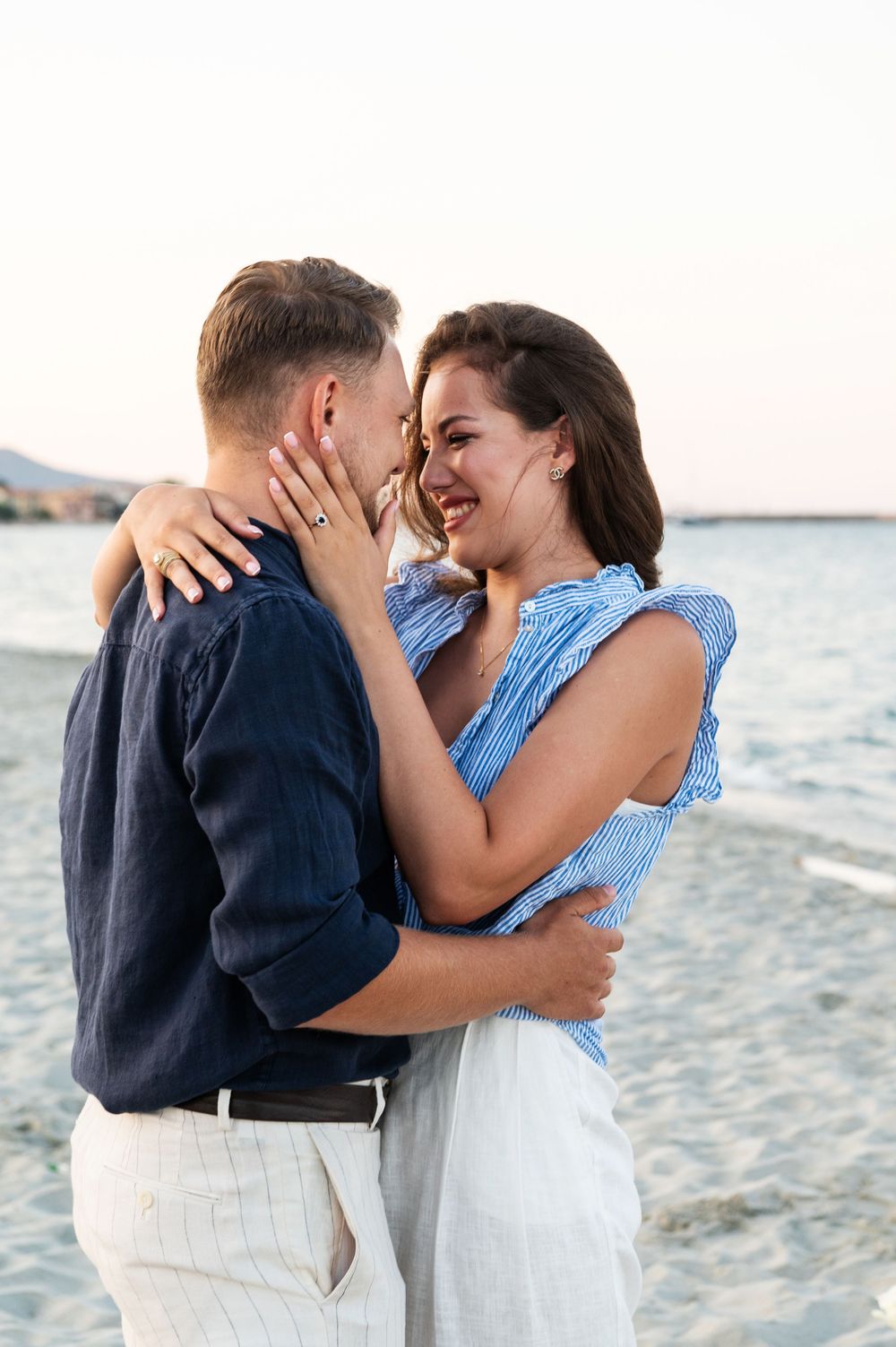 A couple embraces lovingly on a beach at sunset wearing blue and white outfits.