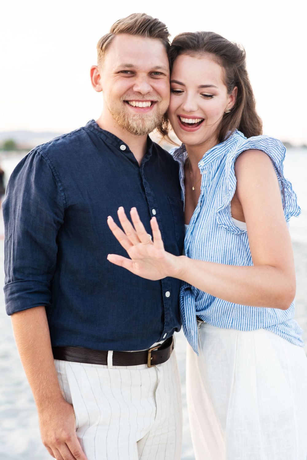 Couple celebrates engagement with laughter and hugs by the ocean.