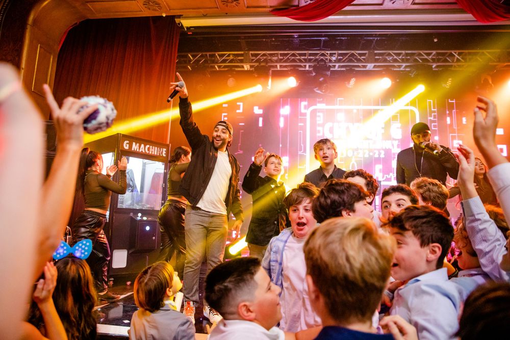 Energetic crowd at a concert venue with bright stage lighting and arms raised in celebration.