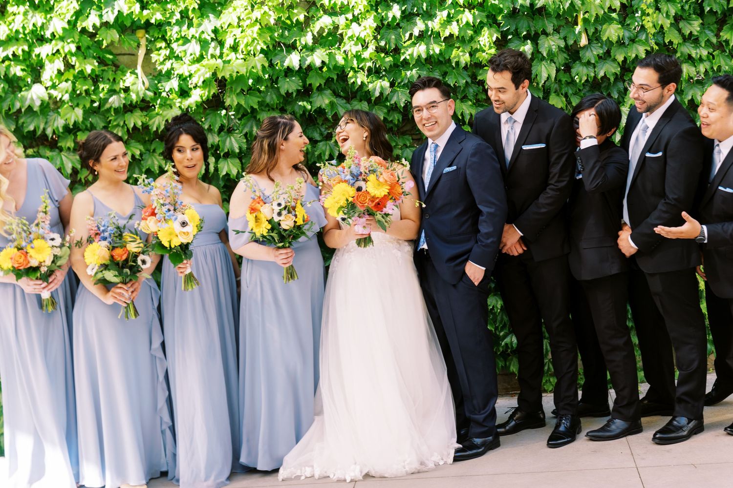 Wedding party in formal attire poses outdoors with bridesmaids in light blue dresses and groomsmen in dark suits.
