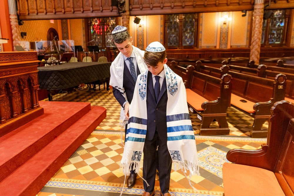 Two people wearing prayer shawls and formal attire stand in a synagogue sanctuary with wooden pews and decorative walls.