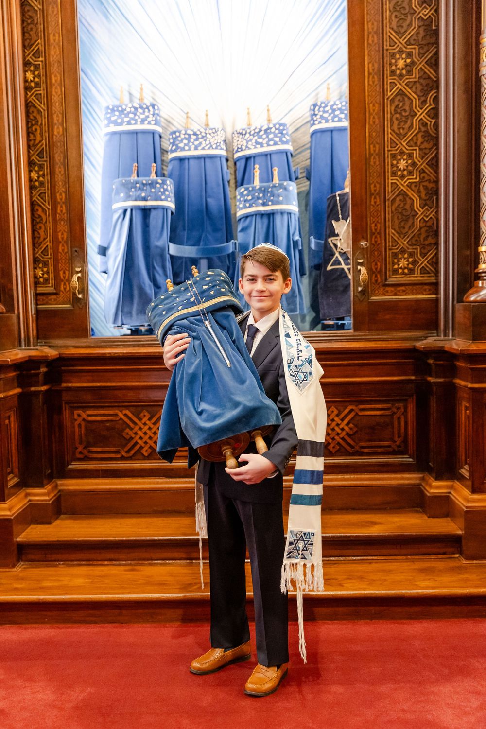 A young person wearing a tallit prayer shawl stands in front of ornate wooden synagogue panels.