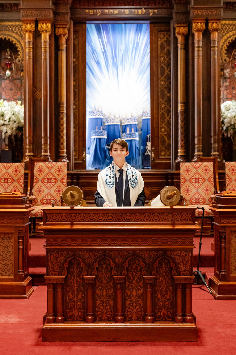 A bar mitzvah ceremony photographed at an elegant synagogue altar with ornate wooden carvings and blue backdrop.