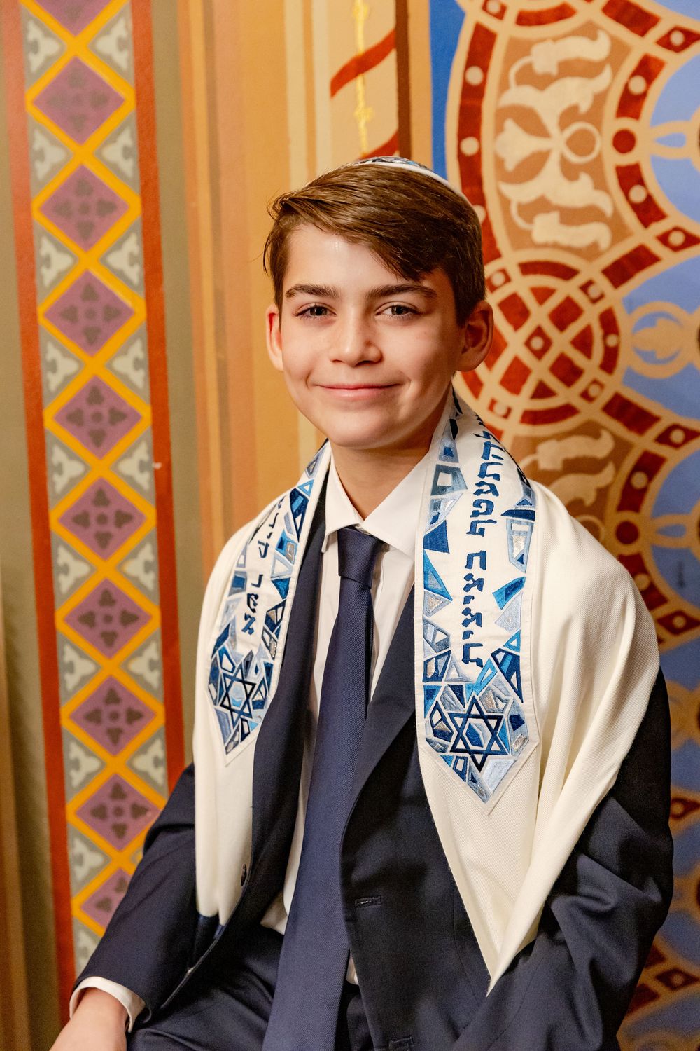 Young man wearing a navy suit and prayer shawl with blue embroidery stands against a decorative patterned wall.
