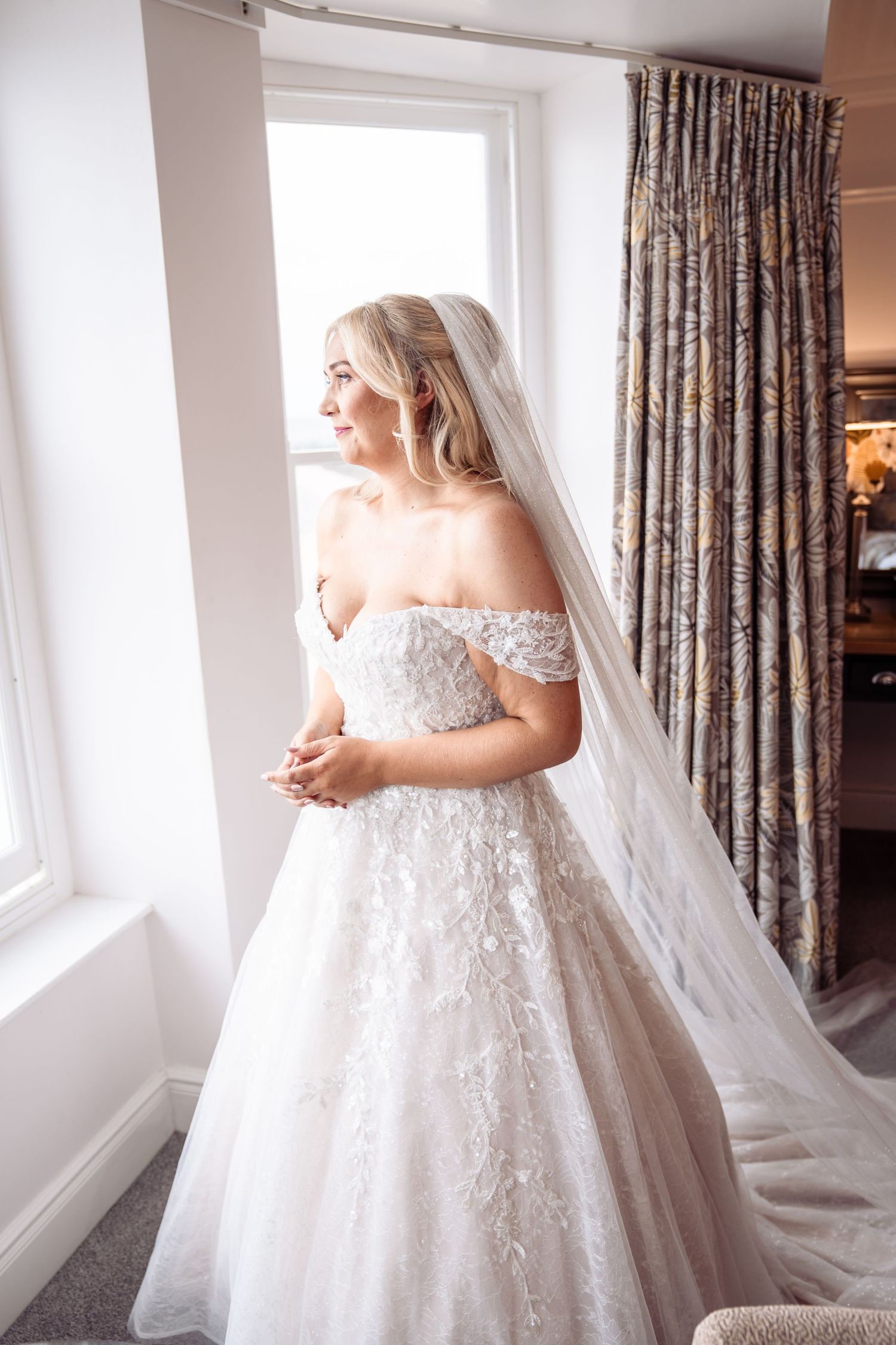 A bride in a stunning off-shoulder lace ball gown twirls by a bright window, her veil flowing elegantly behind her.