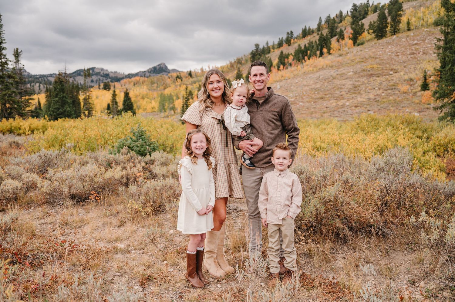 A group of people pose for photos in a scenic mountain meadow during autumn with golden grass and evergreen trees.