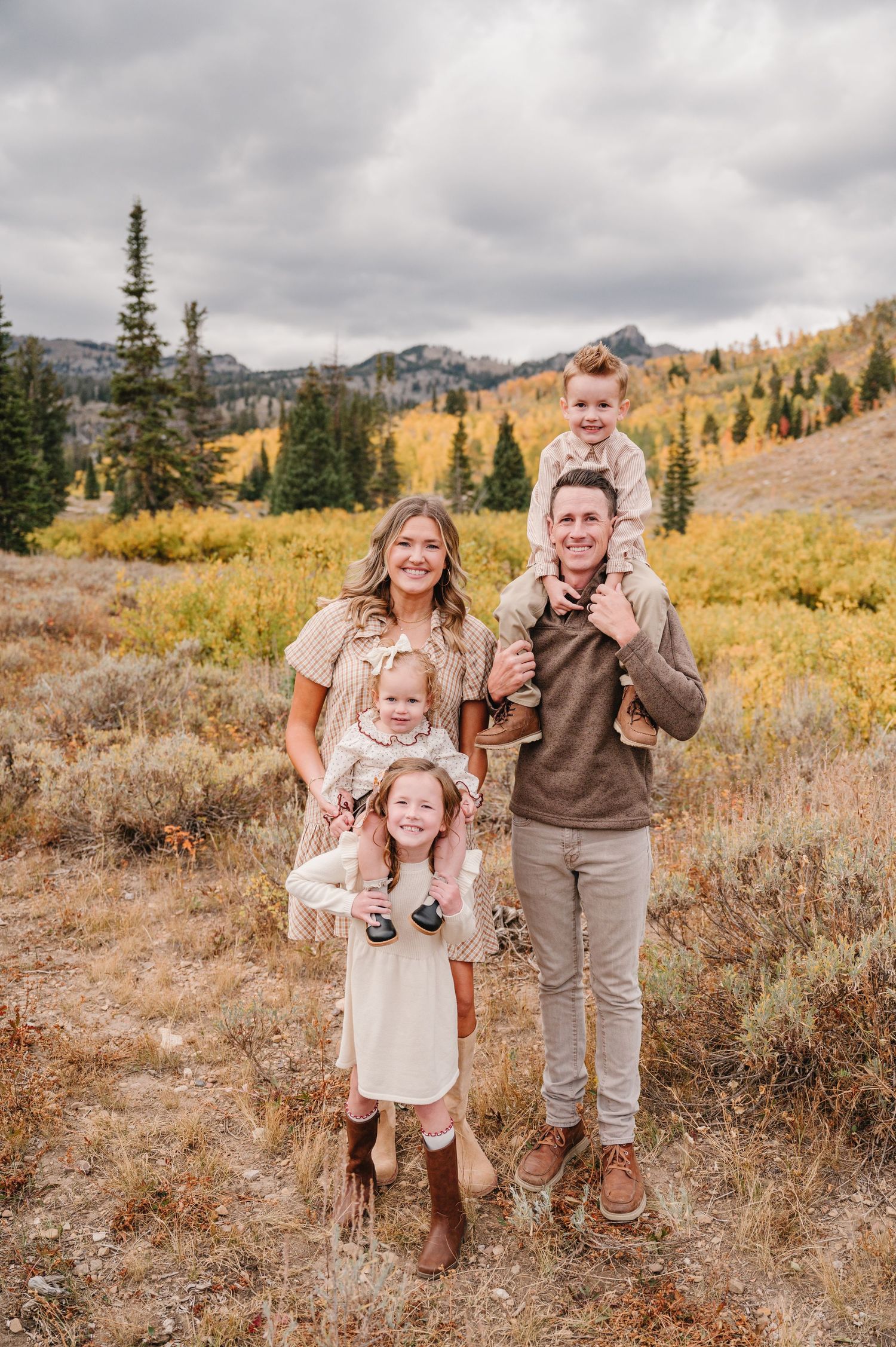 Family in coordinated cream and brown outfits pose together in autumn mountain meadow with golden foliage.