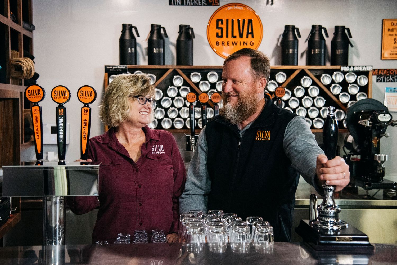 A man and a woman smile at each other behind the bar of a brewery called Silva Brewing.