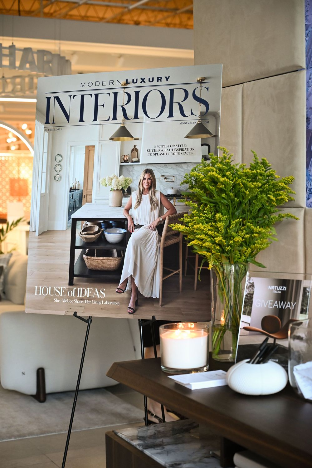 Modern luxury interiors magazine displayed on an easel next to a wooden table with decorative vase and candles.