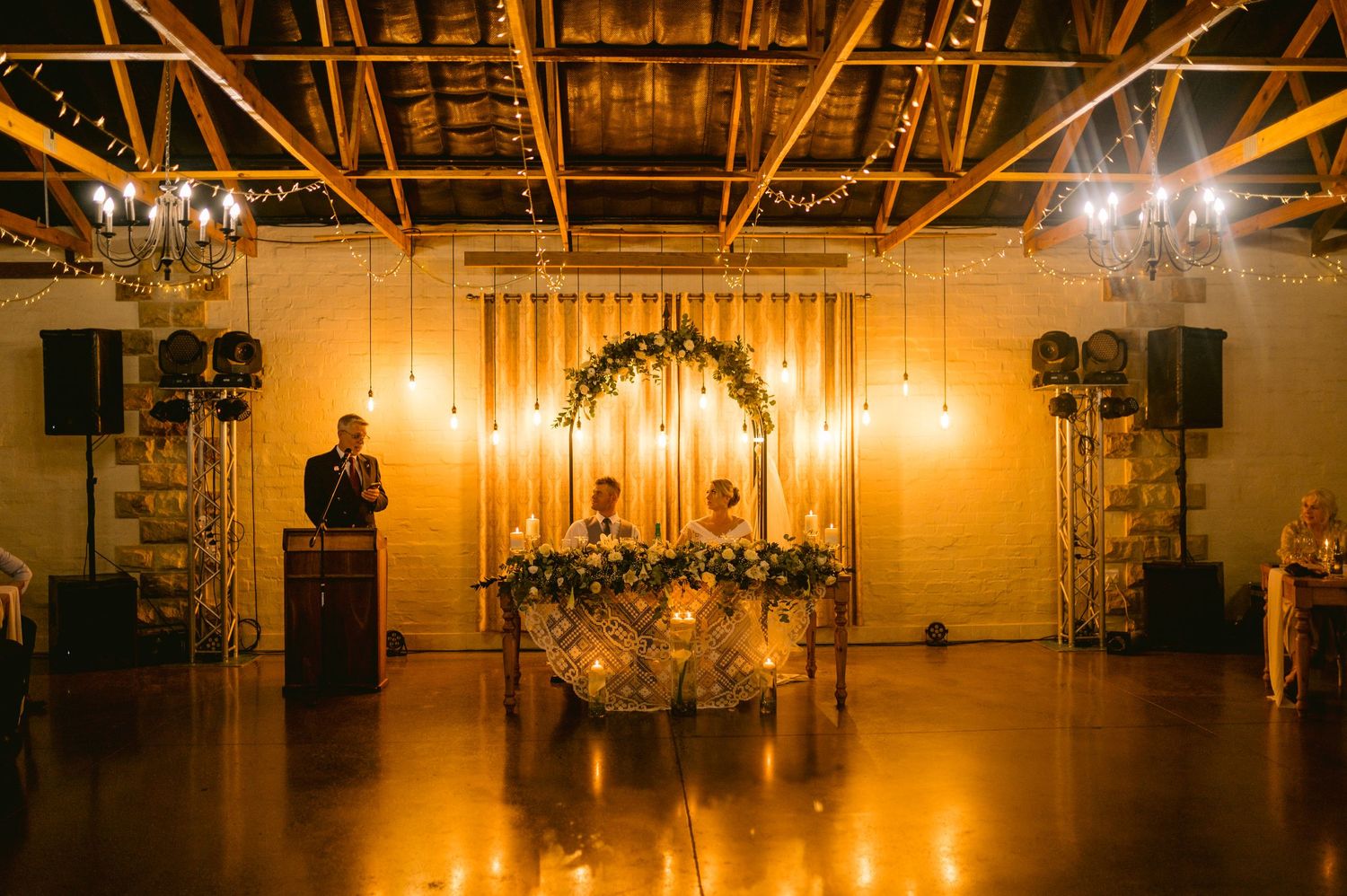 A dimly lit wedding reception venue with warm amber lighting and exposed wooden beams creates an intimate atmosphere.