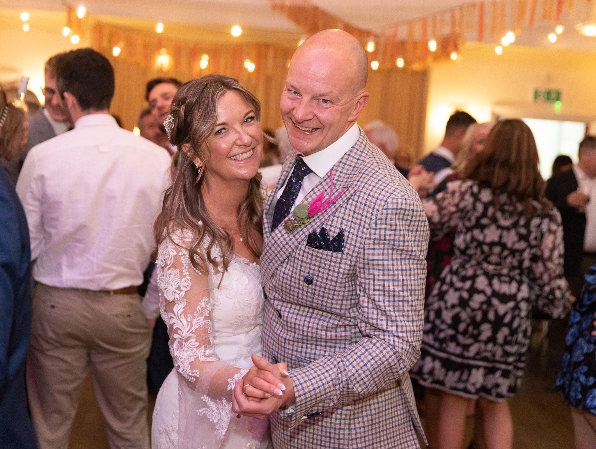 A couple dances together at a wedding reception, with the bride in lace and groom wearing a checkered suit and pink tie.
