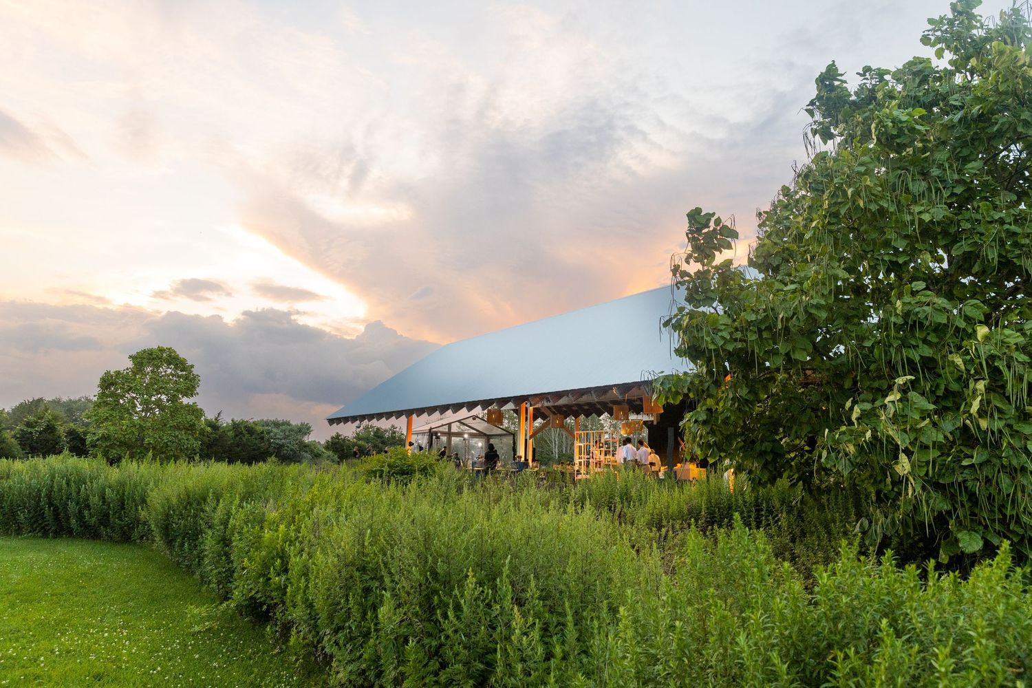 A panoramic view of a lush green field with trees and buildings under a dramatic cloudy sunset sky.